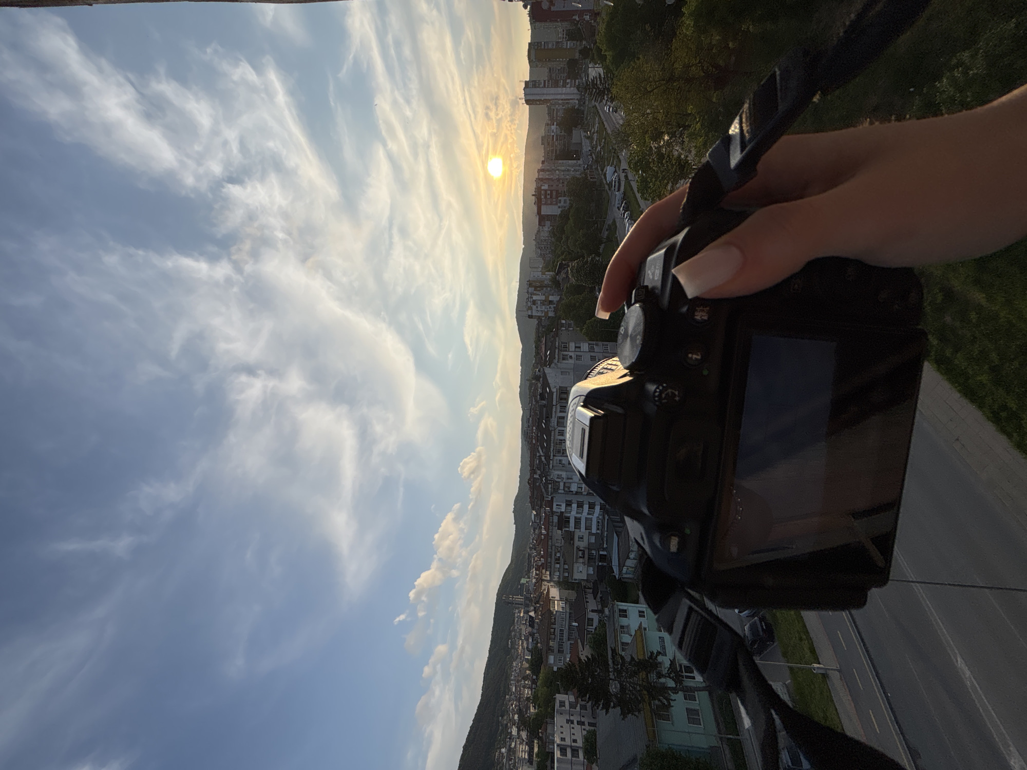 A person is holding a digital single-lens reflex camera, positioned to photograph a cityscape at sunset. The sky is filled with streaks of clouds, and the sun is partially obscured by distant buildings. The city sprawls across a valley, with a forested hill in the background. A road with vehicles is visible in the lower right corner. The camera strap is also visible in the upper right corner.