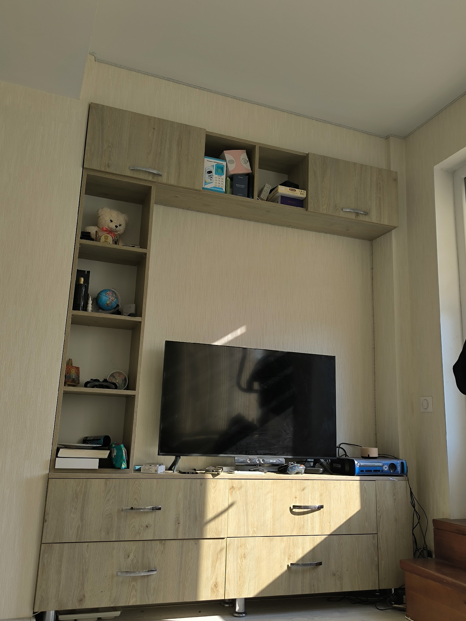 The image depicts a living room scene in Chișinău, Moldova. The focal point is a wall-mounted entertainment unit with a television as the central element. The unit is constructed of light-colored wood-effect material and features overhead cabinets and open shelving. On the shelves, various decorative items and electronics are visible, including a teddy bear, a globe, books, a game controller, and what appears to be a vintage alarm clock. The television is a flat-screen model, positioned on a lower cabinet which also has drawers. Electronic devices, such as a blue console and a black device that resembles a router or modem, are placed on top of the cabinet. The room is illuminated by natural light entering from the right side of the image, casting shadows across the furniture and floor. The walls are covered with textured wallpaper. To the right of the entertainment unit, a portion of a wooden staircase and a light switch are visible. The overall impression is that of a tidy, lived-in space.
