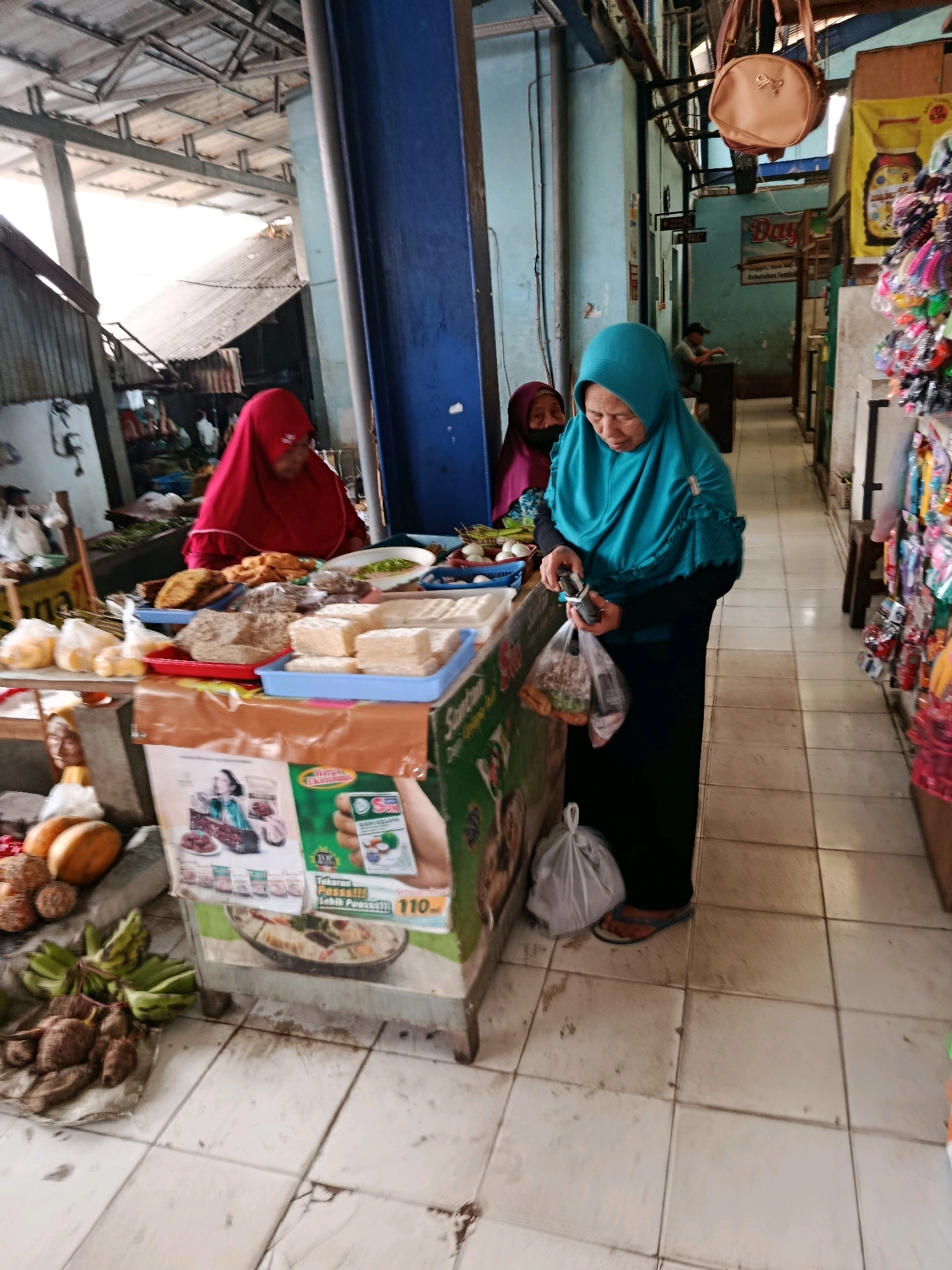 A woman wearing a turquoise hijab and black dress is standing at a market stall in Bululawang, Indonesia. She is holding a plastic bag of goods and looking down at it. In front of her, the stall displays various food items, including blocks of tofu, dried goods, and bananas. Another woman in a red hijab is visible behind the stall. The market appears to be covered, with a corrugated metal roof supported by metal beams. The floor is tiled and shows signs of wear. To the right of the stall, shelves are stocked with colorful packaged items.
