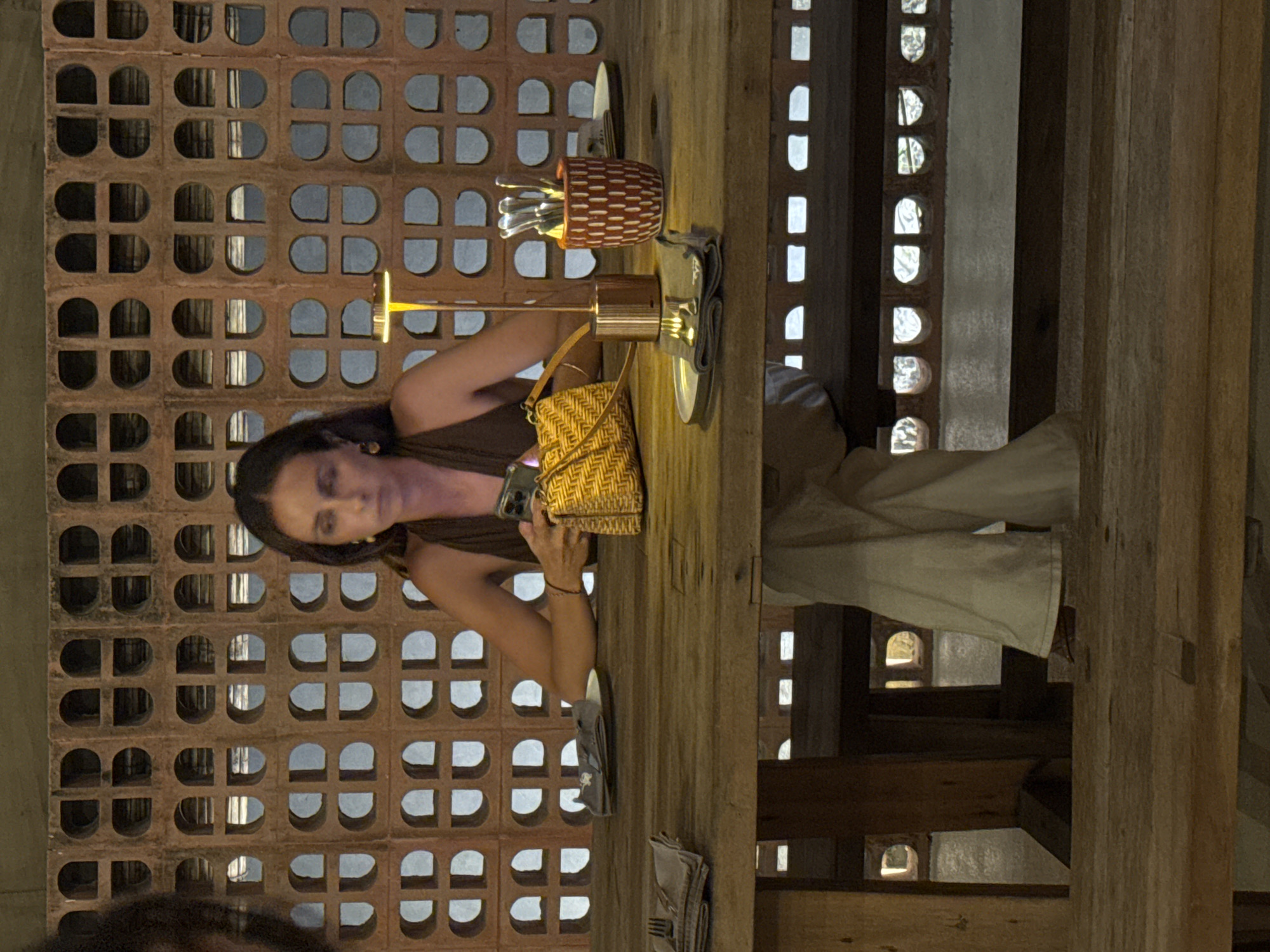 A vertical indoor shot shows a woman seated at a wooden table in what appears to be a restaurant or dining establishment in Kuta Selatan, Indonesia. The woman has dark hair and is wearing a brown tank top. She is looking at a mobile phone in her hands. On the table in front of her are various items, including a woven handbag, utensils, a small lamp, and dining accessories. The background features a decorative brick wall with a grid pattern and an adjacent wooden structure with repetitive arched openings. The lighting is warm, creating an intimate atmosphere.