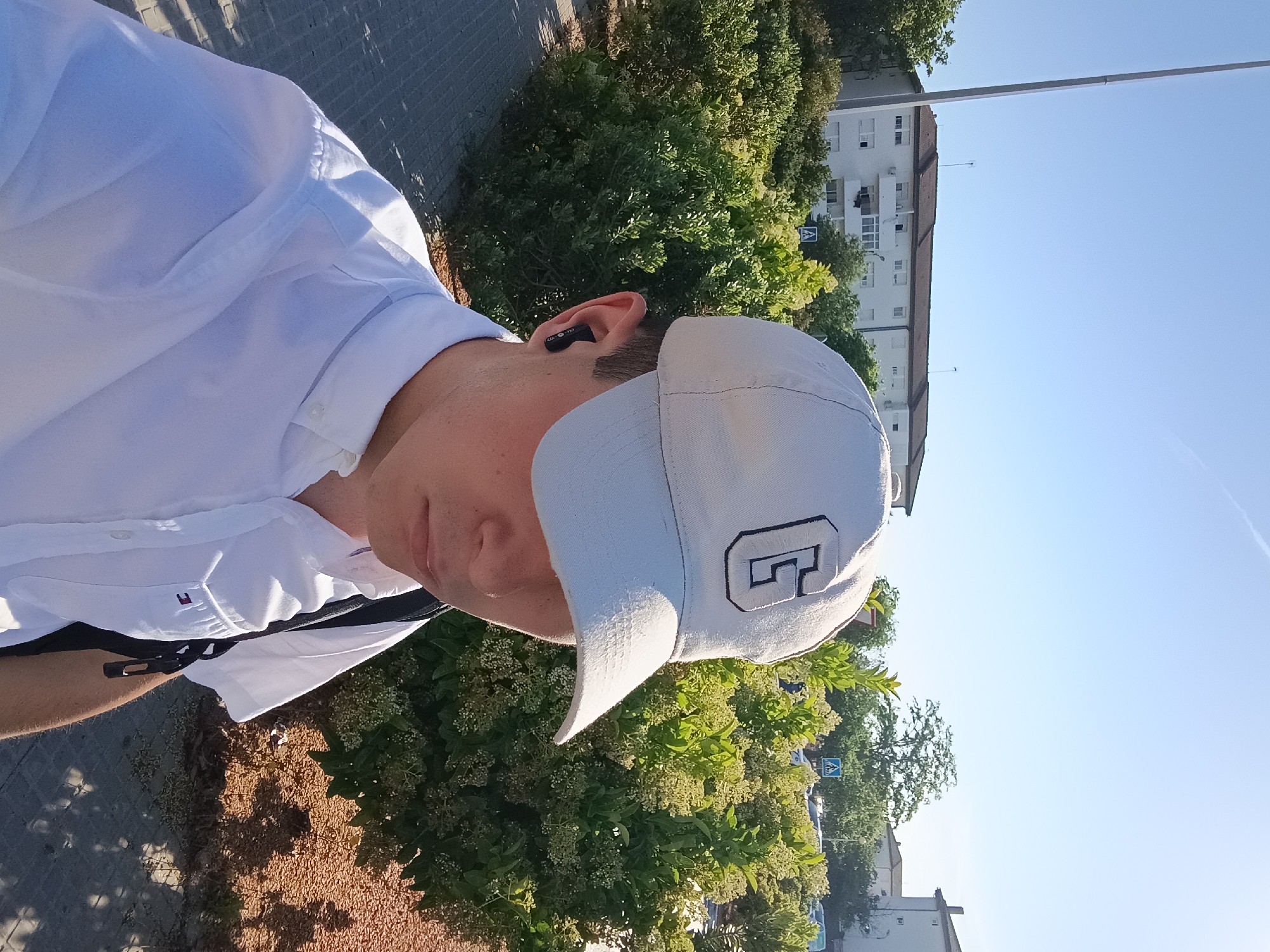 A man wearing a white cap with a stylized letter "C", a white button-down shirt, and possibly a backpack, is standing outdoors near a bush with light yellow flowers and a building in El Port De Sagunt, Spain. He has a wireless earbud in his left ear. The location seems to be a street with a pedestrian crossing sign visible. The ground is partially covered with light brown gravel or small stones. The sky is clear and blue.