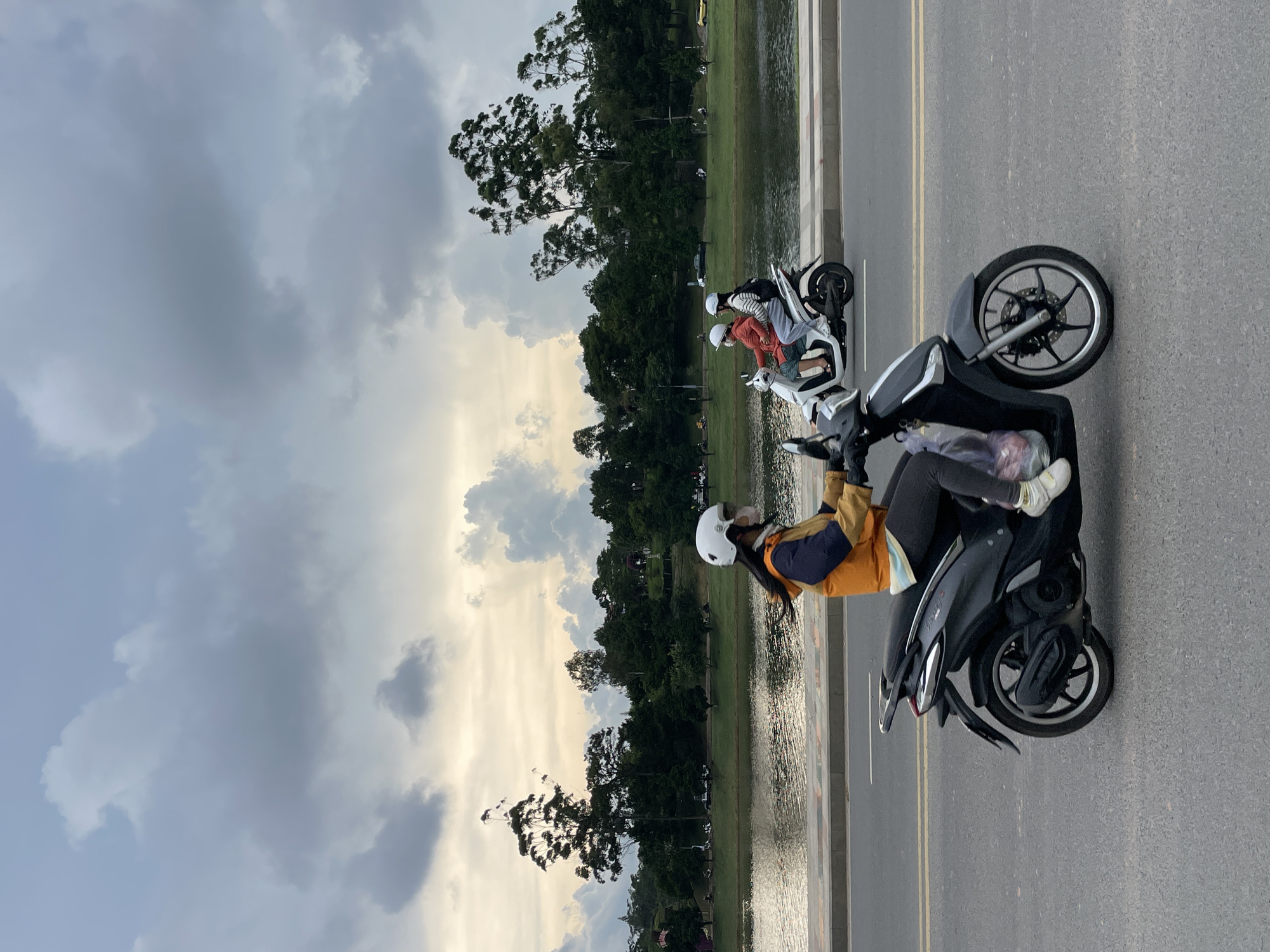 The media file shows a group of people riding motorbikes along a road beside a body of water. The scene is set in Da Lat, Vietnam. The sky is cloudy, suggesting it might be late afternoon or early evening. The weather appears overcast, with some sunlight breaking through the clouds.
In the foreground, a person wearing a yellow and blue jacket and a white helmet is riding a dark-colored motorbike. They are carrying a plastic bag with some items inside. Behind them, another motorbike is visible, carrying two people, also wearing helmets. Further in the background, more people and motorbikes can be seen, as well as trees and a body of water that appears to be a lake or river. The road has yellow lane markings. The overall mood seems relaxed and casual, with people enjoying an outing.