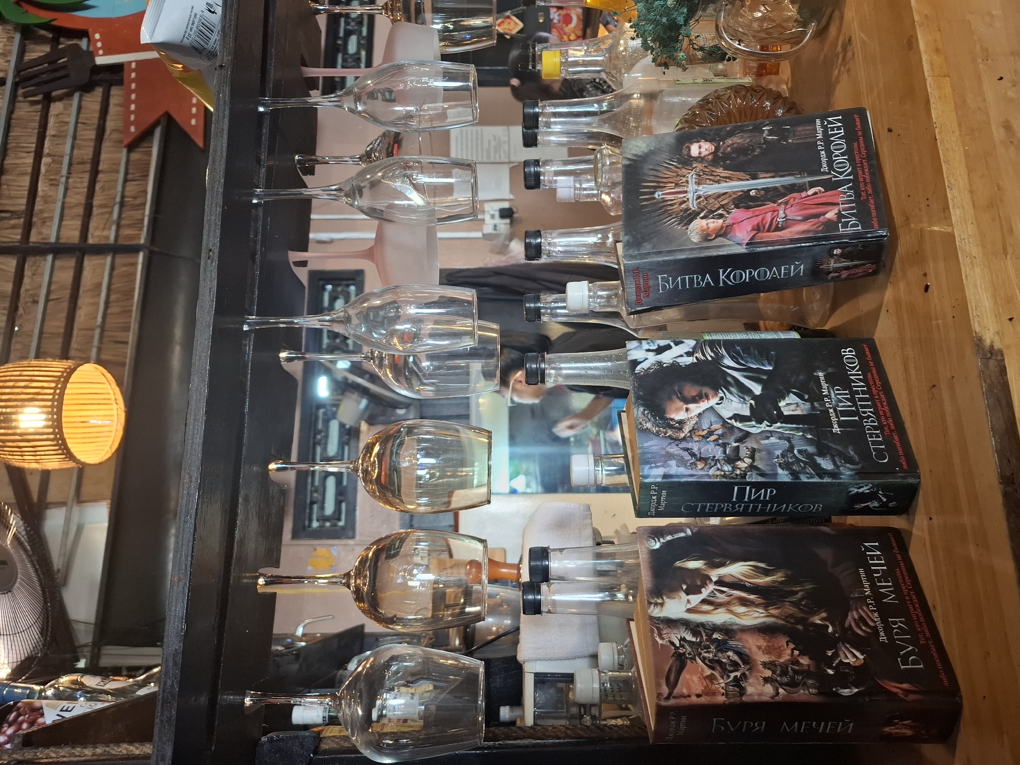 The image shows a wooden bar top in Nha Trang, Vietnam. On the bar are three books by George R. R. Martin. To the left is "A Storm of Swords" in Russian, titled "БУРЯ МЕЧЕЙ". In the middle is "A Feast for Crows" in Russian, titled "ПИР СТЕРВЯТНИКОВ". To the right is "A Clash of Kings" in Russian, titled "БИТВА КОРОЛЕЙ". Behind the books are several glass bottles and glassware hanging from a shelf above. The background appears to be a bar or restaurant interior, with a fan and a lamp visible. No people are clearly identifiable.