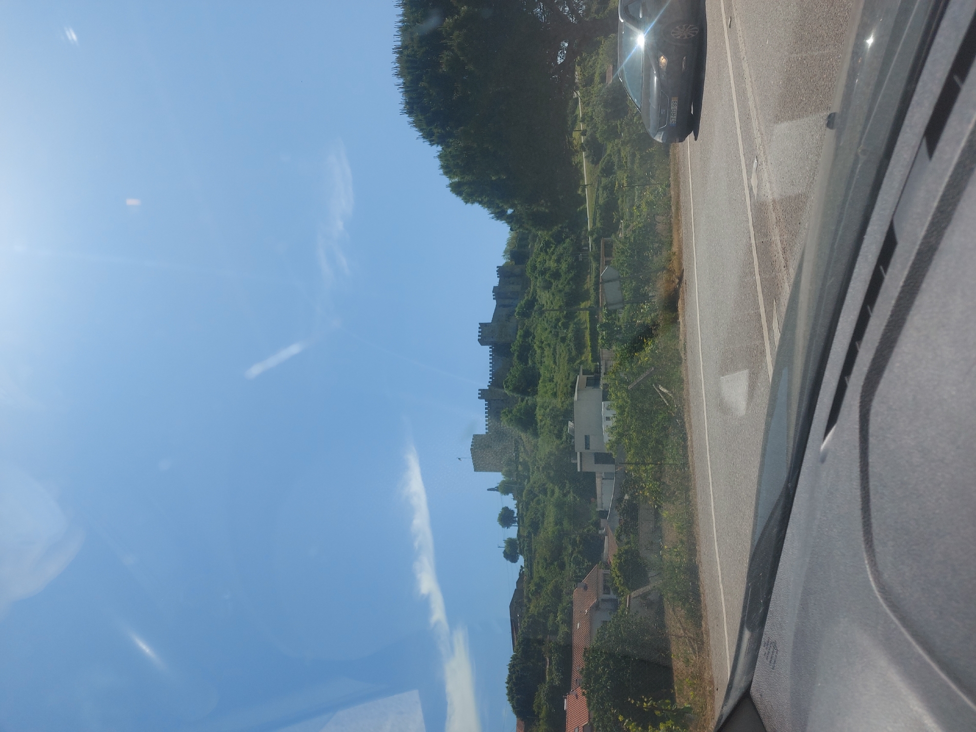 This image presents a bright, sunny daytime view from the passenger side of a moving vehicle, looking out at a scenic landscape featuring a historic castle. The perspective is outward and slightly forward from the car.
The upper portion of the frame is a clear, vibrant blue sky, dotted with a few thin, wispy white clouds and a distinct contrail. A strong lens flare from the sun is visible in the upper left, indicating direct and intense sunlight.
In the middle ground, prominently featured on a verdant hillside, is a large stone castle, characterized by its sturdy walls and battlements. This is likely the Montemor-o-Velho Castle, given the location context. The castle is surrounded by abundant green trees and foliage. Below the castle, nestled into the hillside, are several houses with traditional tiled roofs, indicating a small settlement.
To the right of the scene, a paved road stretches ahead, with a clear white line marking its edge. A dark-colored sedan is visible on the road in front of the viewer's car, its front portion reflecting the bright sunlight. The license plate is partially visible, appearing to read "57AR69," which is consistent with Portuguese vehicle registration plates, reinforcing the specified location of Montemor-o-Velho, Portugal. The immediate foreground on the far right shows the textured grey interior of the car's window frame and door pillar.
The overall atmosphere is one of serene travel through a historic and picturesque landscape on a clear and beautiful day. No people are visible in the image.