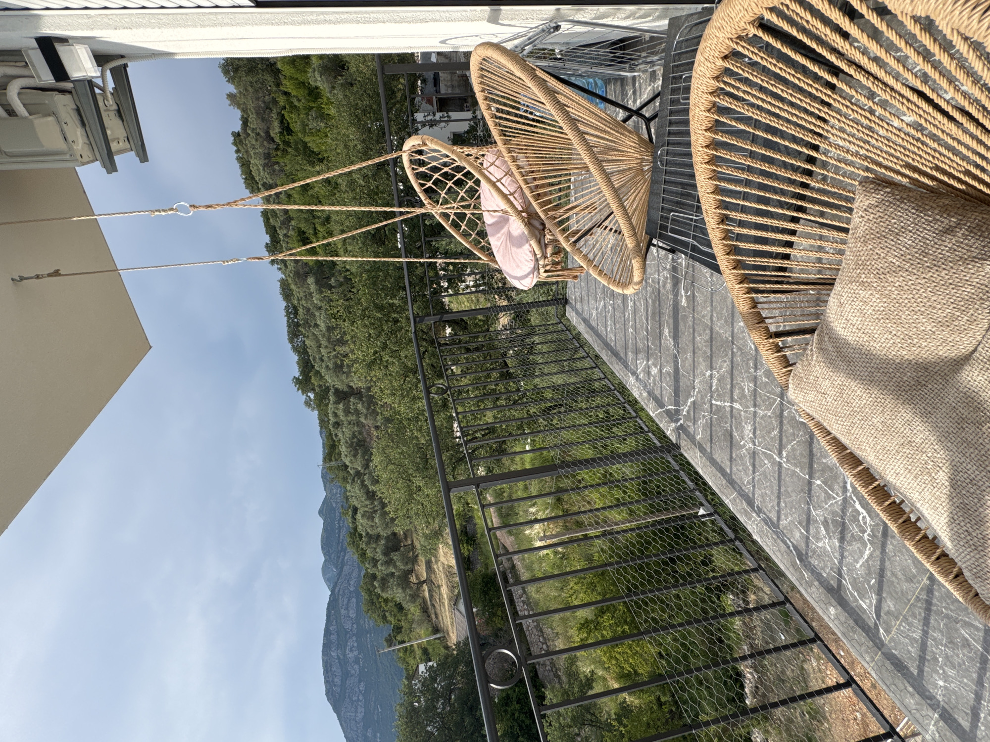 The image depicts a sunlit balcony with two hanging chairs. The chairs are made of woven rope, one with a light pink cushion and the other with a beige cushion. The balcony has a dark, marble-patterned floor and a black metal railing with a wire mesh infill. Beyond the railing, a lush green hillside slopes down, dotted with trees and evidence of human habitation. In the distance, rugged mountains rise against a clear, pale blue sky. The scene suggests a peaceful outdoor setting, possibly a vacation rental or a private residence, with a focus on relaxation and enjoying the natural landscape. The time of day appears to be late morning or early afternoon, given the bright sunlight and clear sky. There is no visible text in the image.