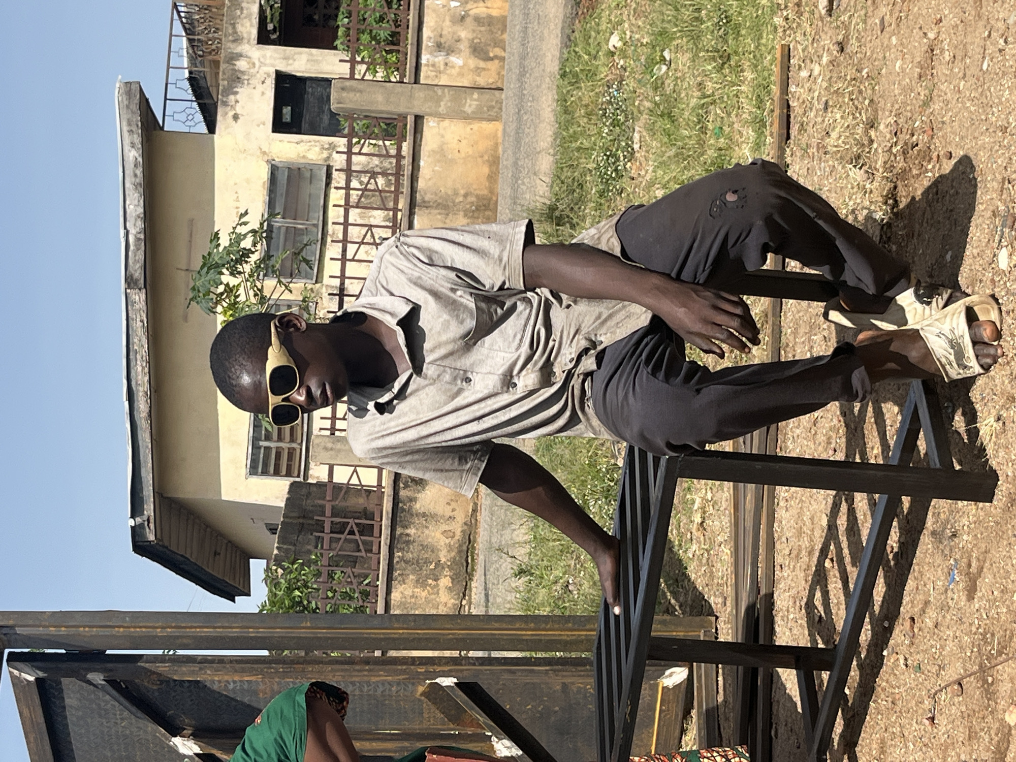 A young Black male with dark skin and short black hair, wearing tan-rimmed sunglasses, a short-sleeved grey collared shirt, and dark grey pants, sits on a black metal bench in an outdoor setting. The bench appears to be part of a metal fabrication workshop or construction site, as indicated by other metal pieces on the ground. The background features a light yellow building with a balcony and a rusty metal fence, typical of residential areas in Jos, Nigeria. The ground is covered in gravel and dry grass. Sunlight casts shadows, suggesting it is daytime. The man is looking towards the right of the frame. Another person, wearing a green shirt, is partially visible on the left edge of the image.