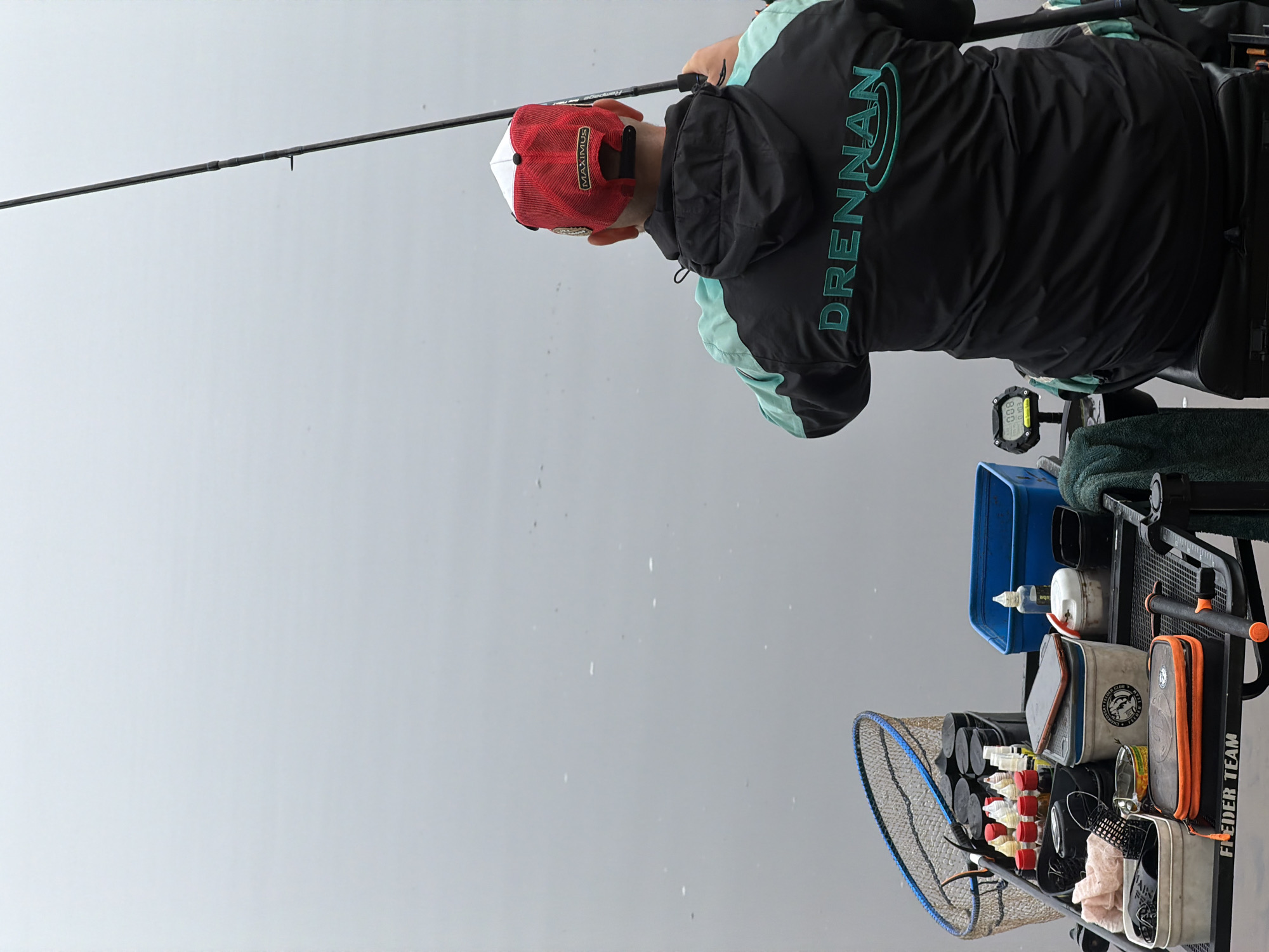 A person is captured from behind, engaged in fishing on what appears to be an overcast or misty day in Noviy, Russia. The image is oriented vertically (originally rotated). The main subject wears a black jacket with mint green accents, featuring the brand "DRENNAN" prominently in mint green text on the back. They also wear a red and white mesh cap with "MAXIMUS" written in white on the red part.
The individual holds a fishing rod, partially obscuring the branding "Rampage." To their right, a comprehensive fishing station is set up. This station includes a blue container, various small white bottles, a grey and white bait box with a circular logo that includes the word "CLUB" and "FEEDER," and an orange-trimmed pouch. A black and orange bar across the front of the station reads "FEEDER TEAM." A digital display attached to the setup shows "8.00" and "0.08," possibly indicating a timer or scale reading. A large fishing net with blue trim rests on the left side of the station.
The background is a uniform light grey, suggesting a heavily overcast sky or a thick layer of fog or mist over a body of water. Small white specks are visible against this grey backdrop, indicative of light rain or falling mist. The overall scene suggests a dedicated angler preparing or actively fishing amidst calm, subdued weather conditions.