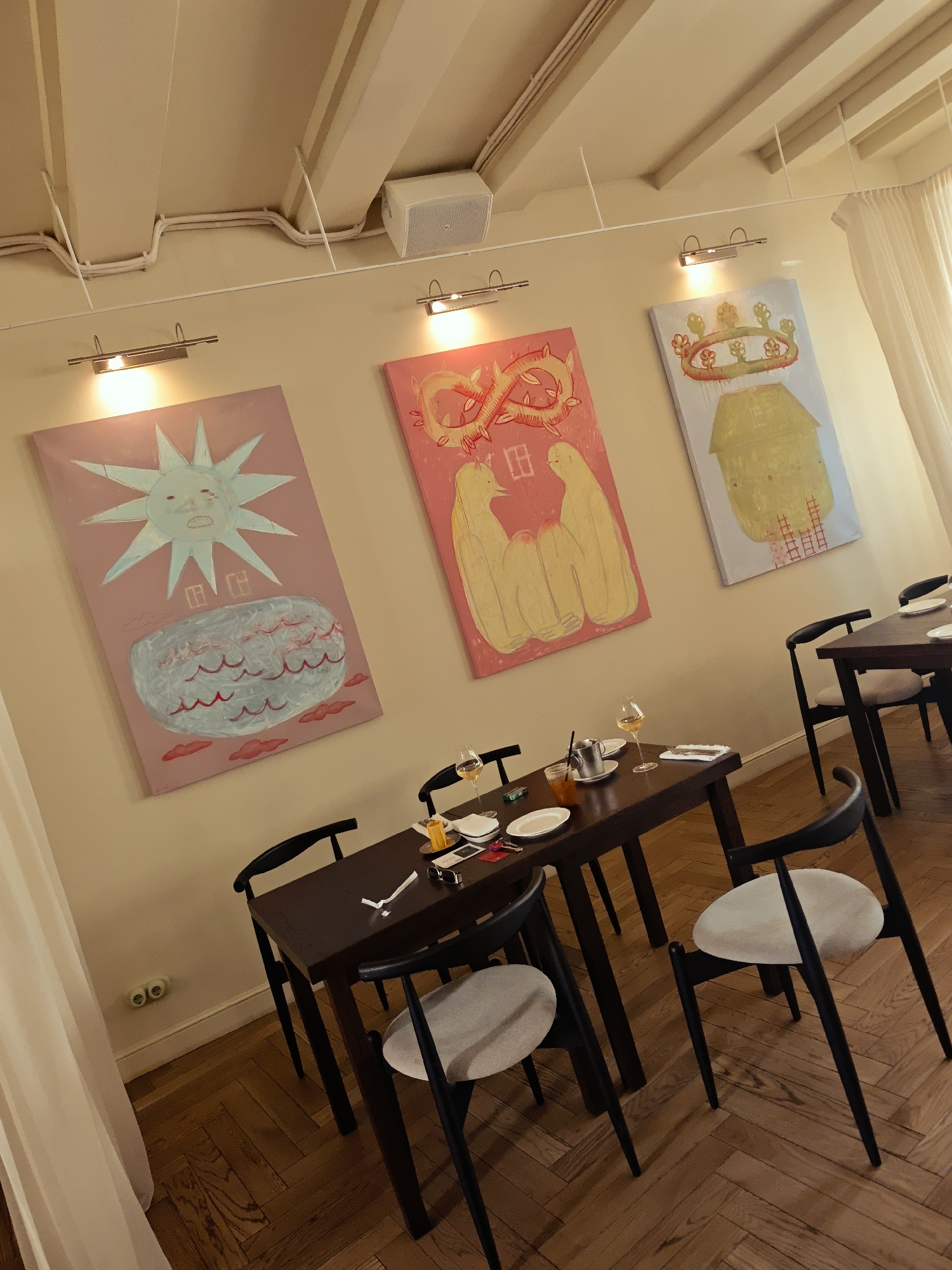 The image shows a dining area in a restaurant with three modern abstract paintings on a light-colored wall. The paintings feature whimsical designs with bold colors. In the foreground, a dark wooden table is set with plates, glasses, and a drink. Several black-and-white chairs with light-colored cushions surround the table. The floor is made of polished wooden planks laid in a herringbone pattern. The room is lit by overhead lights and spotlights illuminating the paintings. The overall impression is one of a contemporary and artistic dining space. There are no people visible in the image.