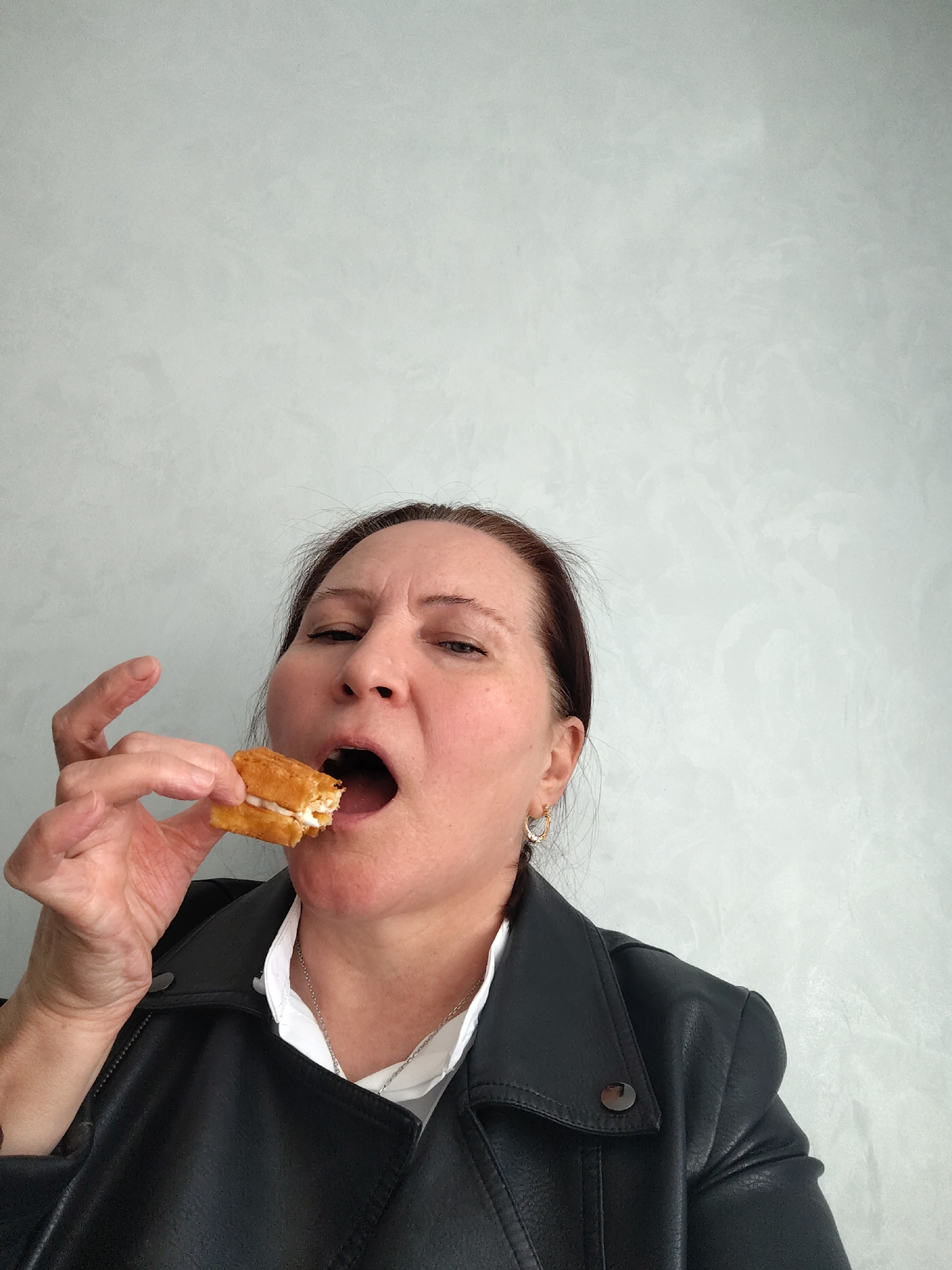 A medium-shot photograph shows a woman about to take a bite of a small, rectangular cake with white filling. The woman has brown hair pulled back, fair skin, and is wearing a black leather jacket over a white shirt with a silver necklace. She is looking towards the camera with her mouth open, holding the cake in her right hand. The background is a light gray wall with a textured finish. There are no obvious indications of the location in the city of Neftekamsk, Russia.