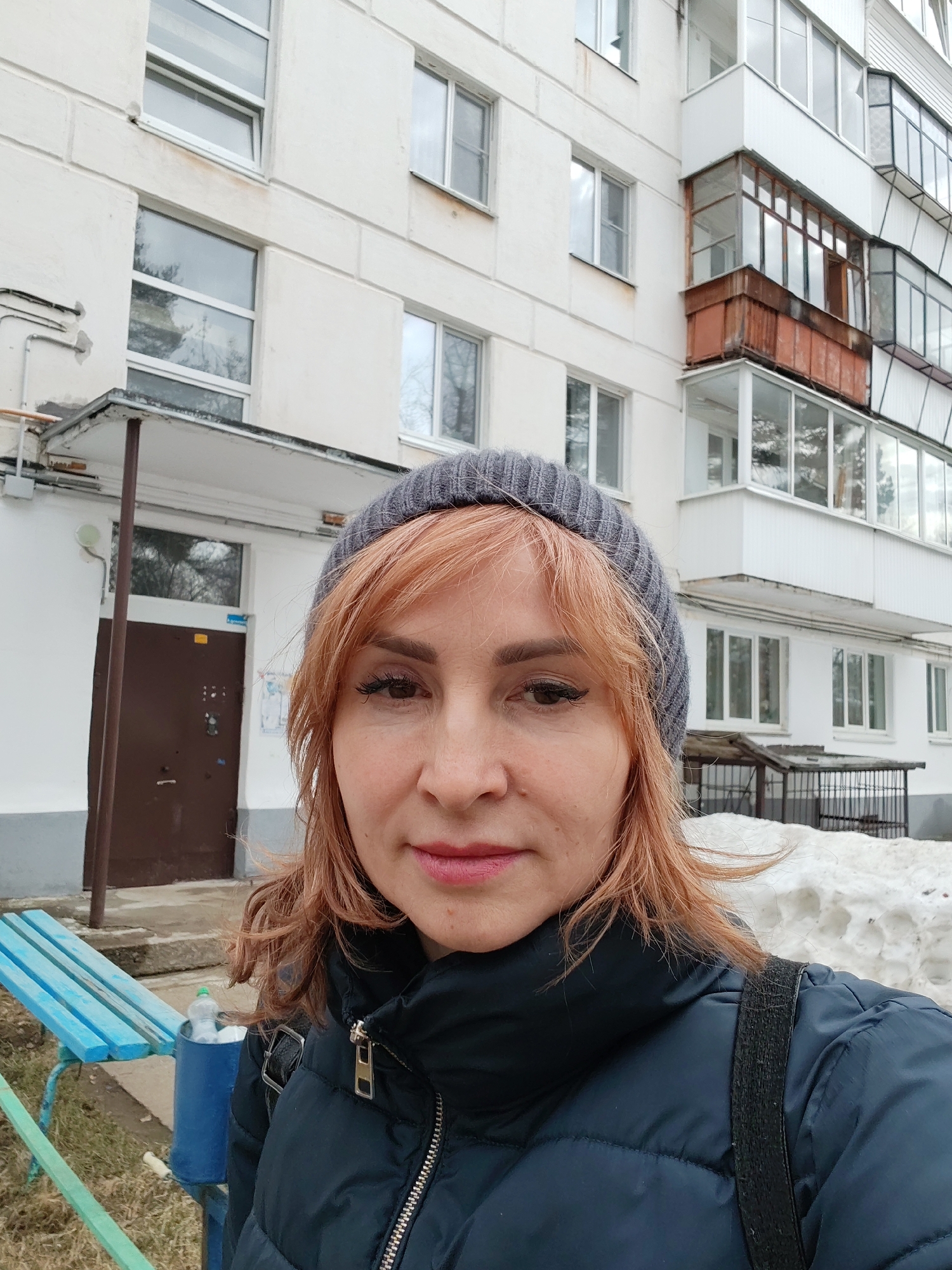 The image shows a woman in a gray beanie and dark blue puffer jacket standing outside a multi-story building. The building has a facade of white brick with several windows and enclosed balconies. In the background, there is a snow pile, a blue bench, and a brown door. The location is identified as the city of Chaikovsky, Russia.