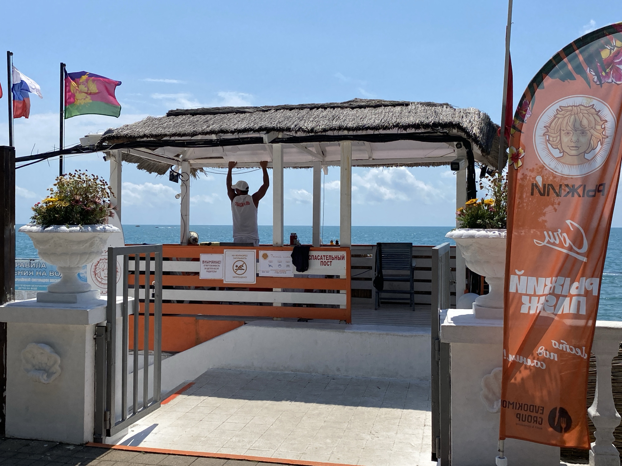 This is a medium shot of a beachside lifeguard station on a sunny day. A lifeguard wearing a white cap and a white tank top with "СПАСАТЕЛЬ" (lifeguard) printed on it stands inside a thatched-roof pavilion, holding onto the roof's support beams. The station is situated near the sea, with the blue water and sky visible in the background. Flags, including the Russian flag and another flag with a purple and green design, fly on poles to the left of the pavilion. An orange promotional flag with an illustration of a person's face and text in an unfamiliar script stands to the right.
Several signs are posted on the pavilion. One sign reads "ВНИМАНИЕ! ОТВЕТСТВЕННОСТЬ ЗА ДЕТЕЙ НЕСУТ РОДИТЕЛИ!" (ATTENTION! PARENTS ARE RESPONSIBLE FOR CHILDREN!). Another sign displays symbols and text, and a larger sign in Russian states "СПАСАТЕЛЬНЫЙ ПОСТ" (LIFEGUARD POST). Decorative white urns with plants are positioned to the left and right of the main walkway leading to the station. The scene suggests a typical summer day at a beach resort in Sochi, Russia. The weather appears to be clear and warm, typical for a beach setting. The overall impression is one of an operational lifeguard station ready to ensure beach safety.