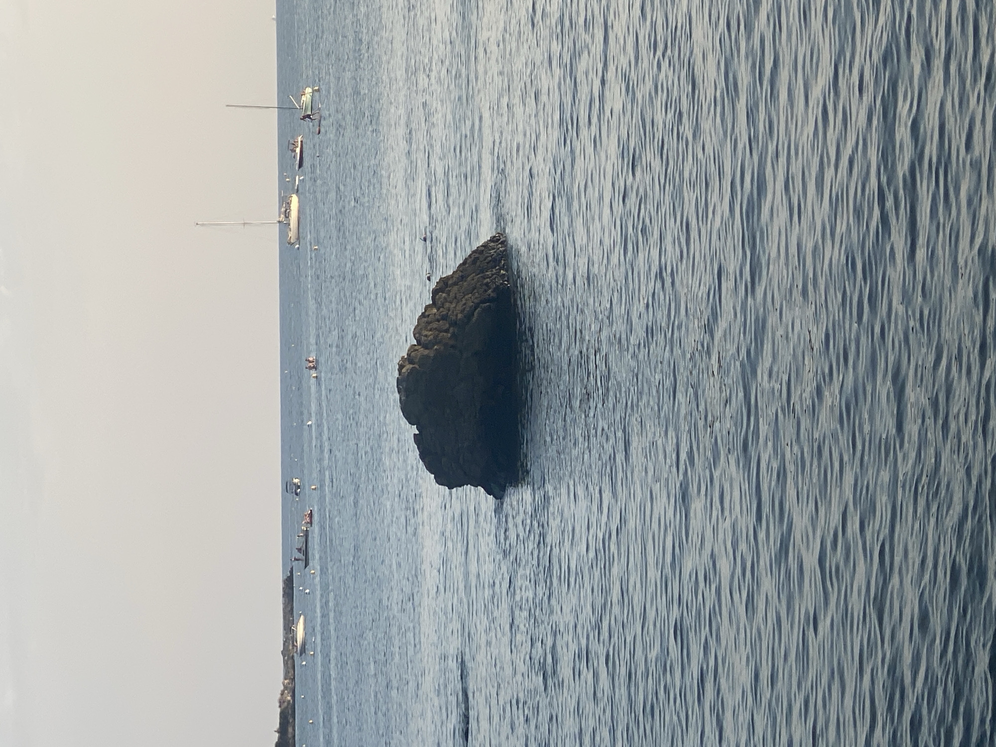 A large, dark rock formation juts out of the water in the foreground of the image. The water is a deep blue and has gentle ripples. In the background, there are several boats and people in the water. Some of the boats are sailboats, while others are smaller motorboats. There are also a few people on paddleboards. The sky is a pale blue with some clouds. The location appears to be a coastal area, likely near Antibes, France, given the presence of sailboats and the overall Mediterranean atmosphere.