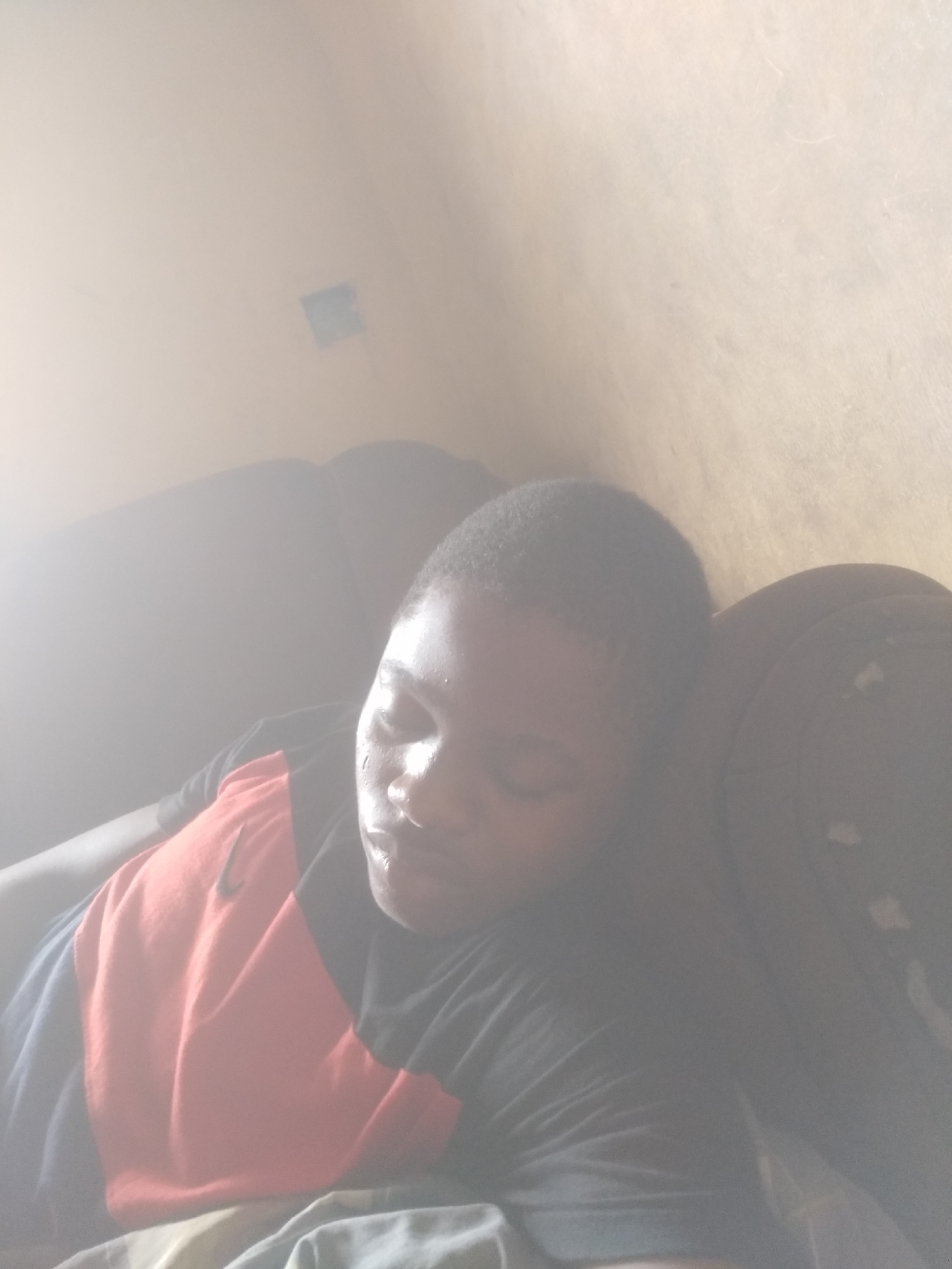 An overexposed, portrait-oriented image shows a person resting or asleep indoors. The subject, likely a young adult or adult with short dark hair, is lying on what appears to be a dark-colored couch or sofa. Their eyes are closed, and their facial expression suggests peaceful sleep or deep rest.
The person is wearing a short-sleeved t-shirt that features a distinctive design with color blocks. The upper chest area is black, transitioning to a prominent orange section across the mid-torso, and a dark blue or black section is visible on the lower left side of the shirt. A black Nike swoosh logo is clearly visible on the orange part of the t-shirt.
A strong, bright light source, possibly sunlight, illuminates the scene from the upper left, causing significant overexposure and a washed-out appearance, particularly on the left side and top of the image. This bright light creates a glare, especially noticeable on the person's face, highlighting what might be sweat or moisture on their skin. The background features a light-colored wall, with a faint, small, dark rectangular object (possibly a light switch or outlet cover) visible on it towards the upper center. The overall impression is one of quiet repose in a brightly lit, likely domestic, setting during the daytime.