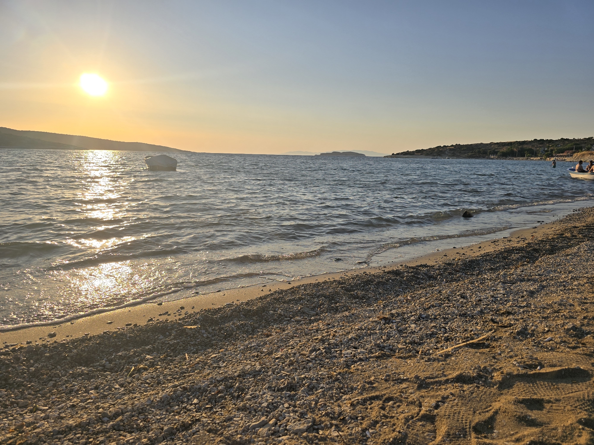 The image depicts a serene sunset scene on a beach in Foça, Turkey. The sun, a bright orb near the horizon, casts a golden glow across the calm sea, creating shimmering reflections on the water's surface. A small, white boat, partially covered, floats gently in the distance. The shoreline is a mix of sand and pebbles, with gentle waves lapping at the shore. In the background, a hilly coastline stretches across the horizon, dotted with sparse vegetation. Towards the right of the frame, a few people are visible near a white boat, some wading in the shallow water. The sky is clear, transitioning from a warm orange near the horizon to a pale blue overhead, indicating the late hour of the day.
