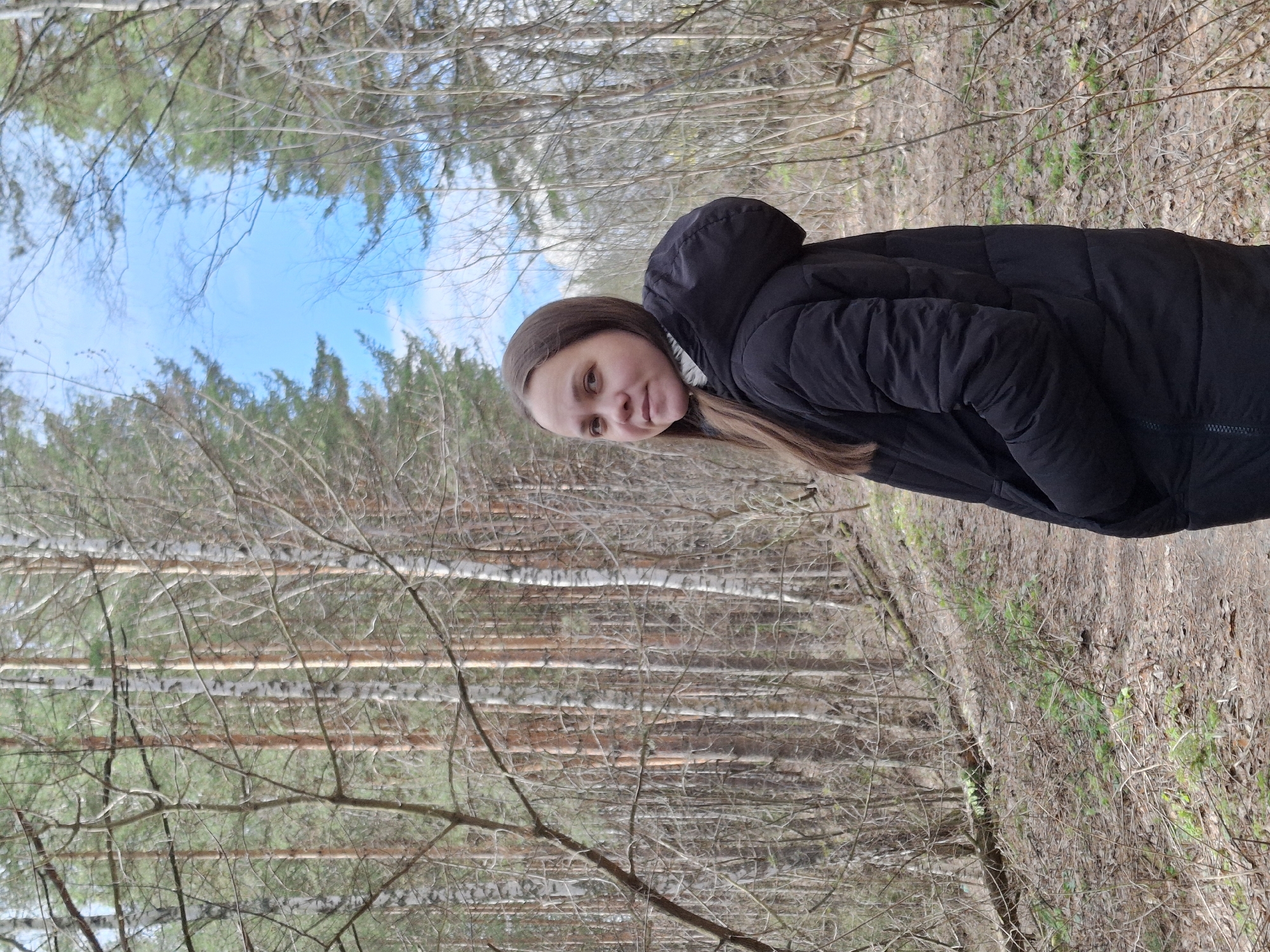 A woman stands in a forest. The location is the city of Chaikovsky, Russia. The woman is wearing a black hooded jacket and has long brown hair. The forest consists of bare trees and a visible blue sky through the branches. The ground is covered with dry leaves and vegetation.