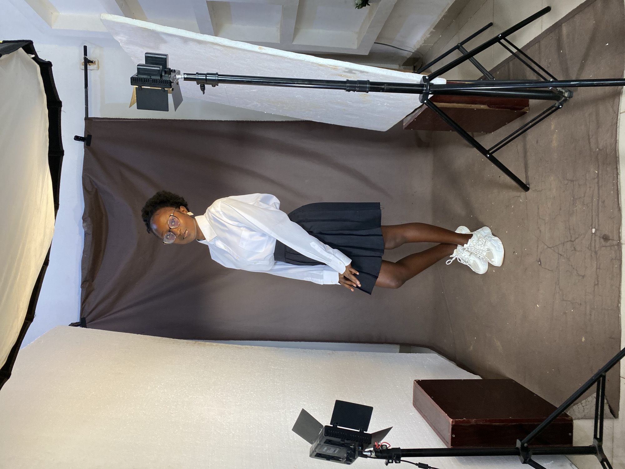 A young Black female is posed in what appears to be a studio setting in Bukuru, Nigeria. She is wearing a white collared shirt, a black pleated skirt, and white sneakers. She has on round glasses and her hair is styled in a short, curly afro. She stands in front of a brown backdrop, with a large white reflector to her right and a photographic light on a stand to her left. Another light on a stand is positioned behind her and to the right, partially obscured by the white reflector. The floor is covered with a brown mat. To the left of the subject, a dark brown wooden box is placed on the floor. The environment suggests a professional photography setup.