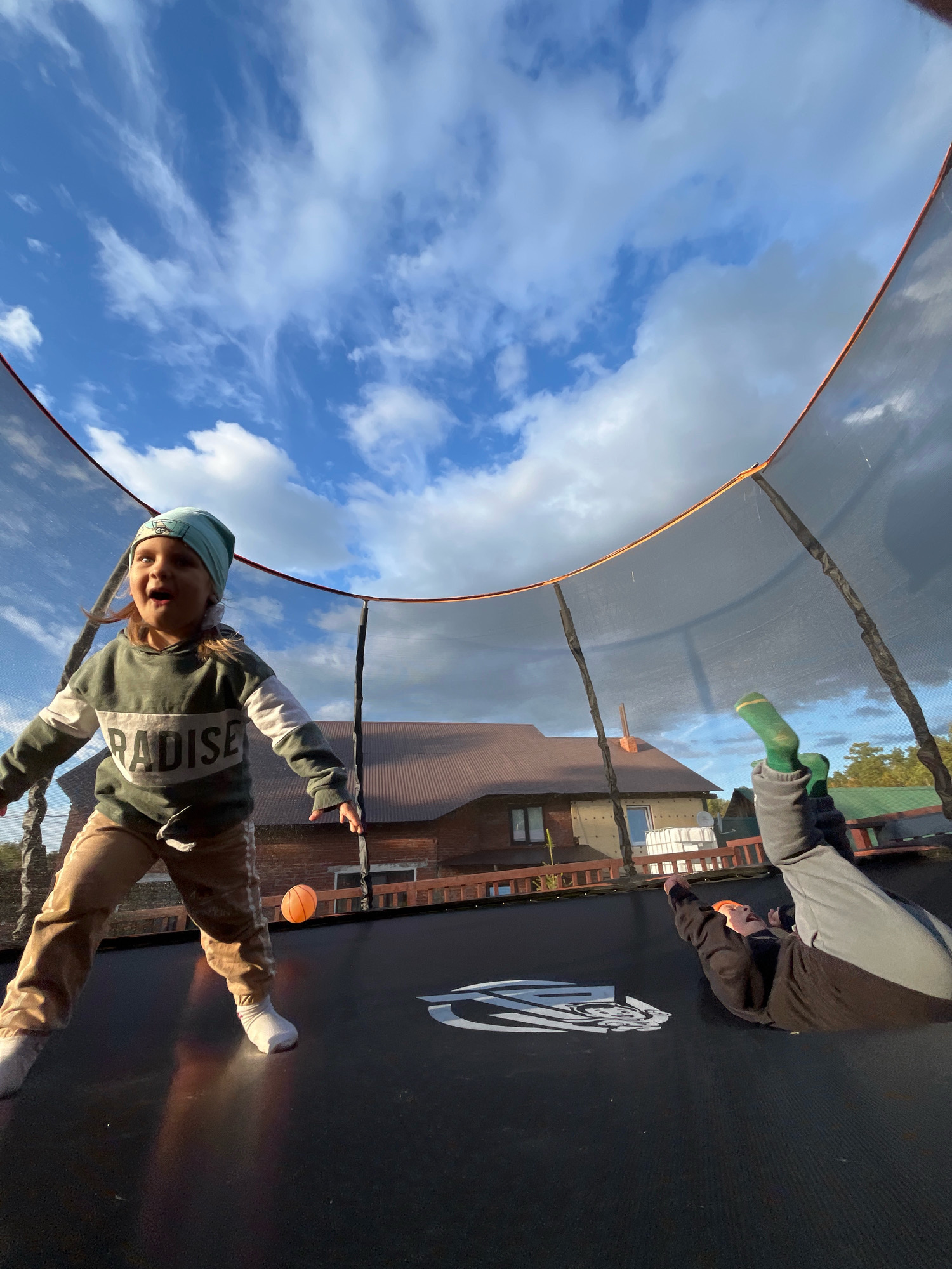 The image shows two children on a trampoline in what appears to be a residential backyard, likely in Chaikovsky, Russia. A young girl, wearing a green and white striped sweatshirt, tan pants, and a blue beanie, is in the foreground, mid-jump and with an open mouth. She is about to kick an orange ball that is suspended in the air. To the right and further back on the trampoline, another child is lying on their back, with their legs extended upwards, wearing a black sweatshirt and grey pants. The trampoline is enclosed by a safety net, and the background shows a house with a brown roof and a clear blue sky with scattered clouds. The perspective is from below, looking up at the children and the sky.