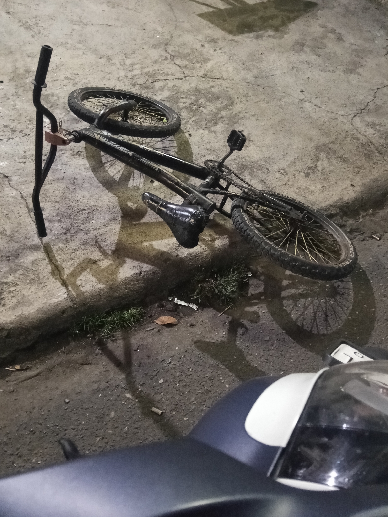 A black BMX bicycle is lying on its side on a cracked concrete surface, likely a sidewalk or road, in Makale, Indonesia. The bicycle is dark in color with gold-colored spokes on the wheels. The handlebars are turned towards the viewer's left, and the seat is pointed upwards. Shadows of the bicycle and possibly other objects are cast on the ground, suggesting it is nighttime or dusk. In the foreground, a portion of a dark-colored vehicle, possibly a scooter or motorcycle, is visible, with a white panel on its front. The surrounding environment appears to be an urban or semi-urban area, with the concrete surface showing signs of wear and tear. There are also patches of grass and some debris on the ground.