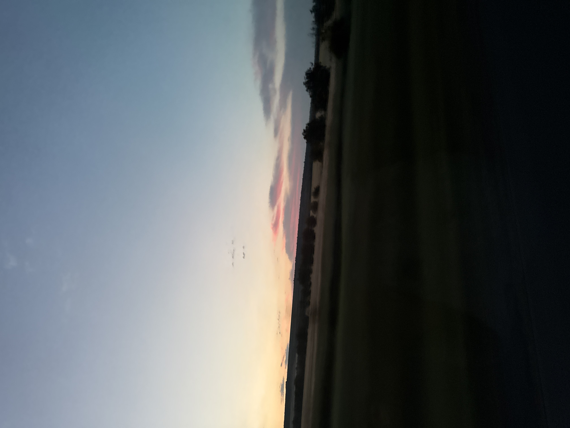 The image depicts a landscape at dusk, with a gradient of colors in the sky ranging from a light blue at the top to warmer hues of pink and orange closer to the horizon. A line of trees is visible along the horizon, silhouetted against the sky. To the right of the trees, a dark, shadowed area suggests a field or a road. The overall mood is serene and peaceful, characteristic of the end of the day. There is no visible text in the image.