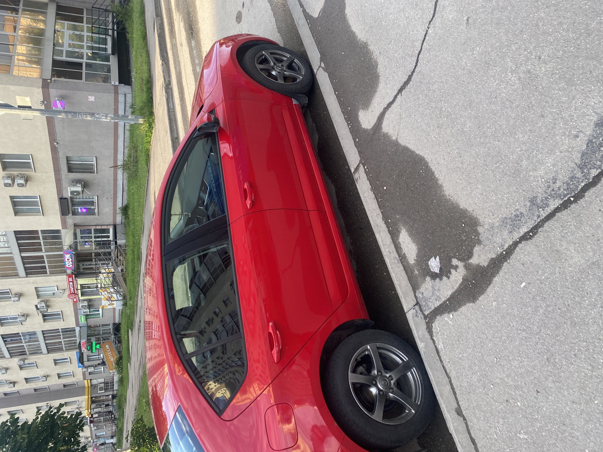 The image shows a bright red sedan parked on the side of a paved street in front of a multi-story building. The building has a light-colored facade with numerous windows and air conditioning units. Several storefronts are visible at the ground level, including one with an "OZON" sign and another with a green cross, suggesting a pharmacy. The street has a concrete curb and shows some water stains and reflections. The car's doors are closed, and no people are visible inside or immediately around the vehicle. The overall scene depicts a typical urban environment in Novokuybyshevsk, Russia.