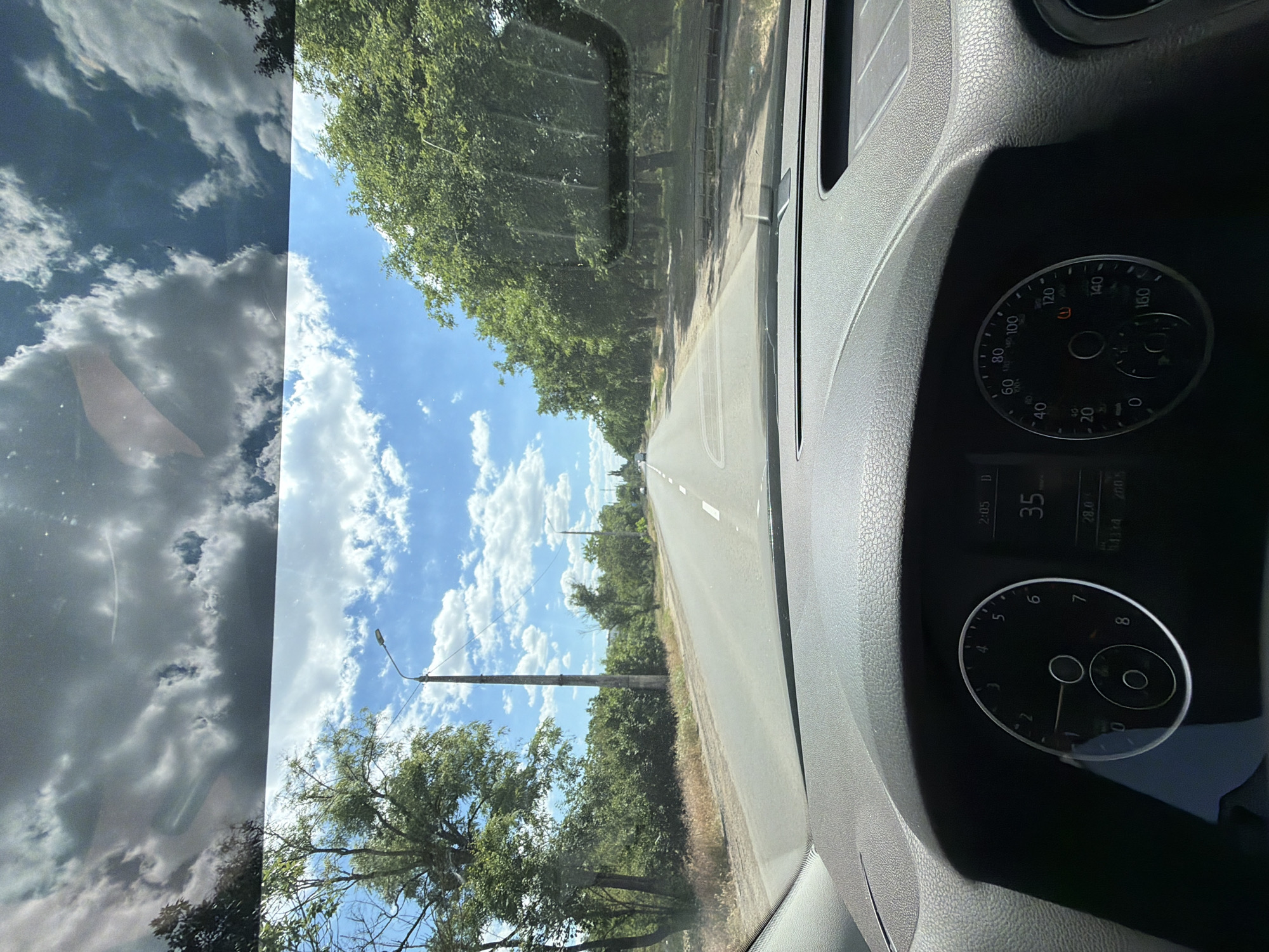 The image is taken from the interior of a vehicle, looking forward through the windshield. The dashboard is visible in the foreground, with a speedometer and other gauges. The road ahead is paved and appears to be a two-lane road lined with trees and vegetation. The sky is blue with scattered white clouds, and there are also reflections on the windshield. The overall scene suggests a journey on a road outside of a city, under a partly cloudy sky.