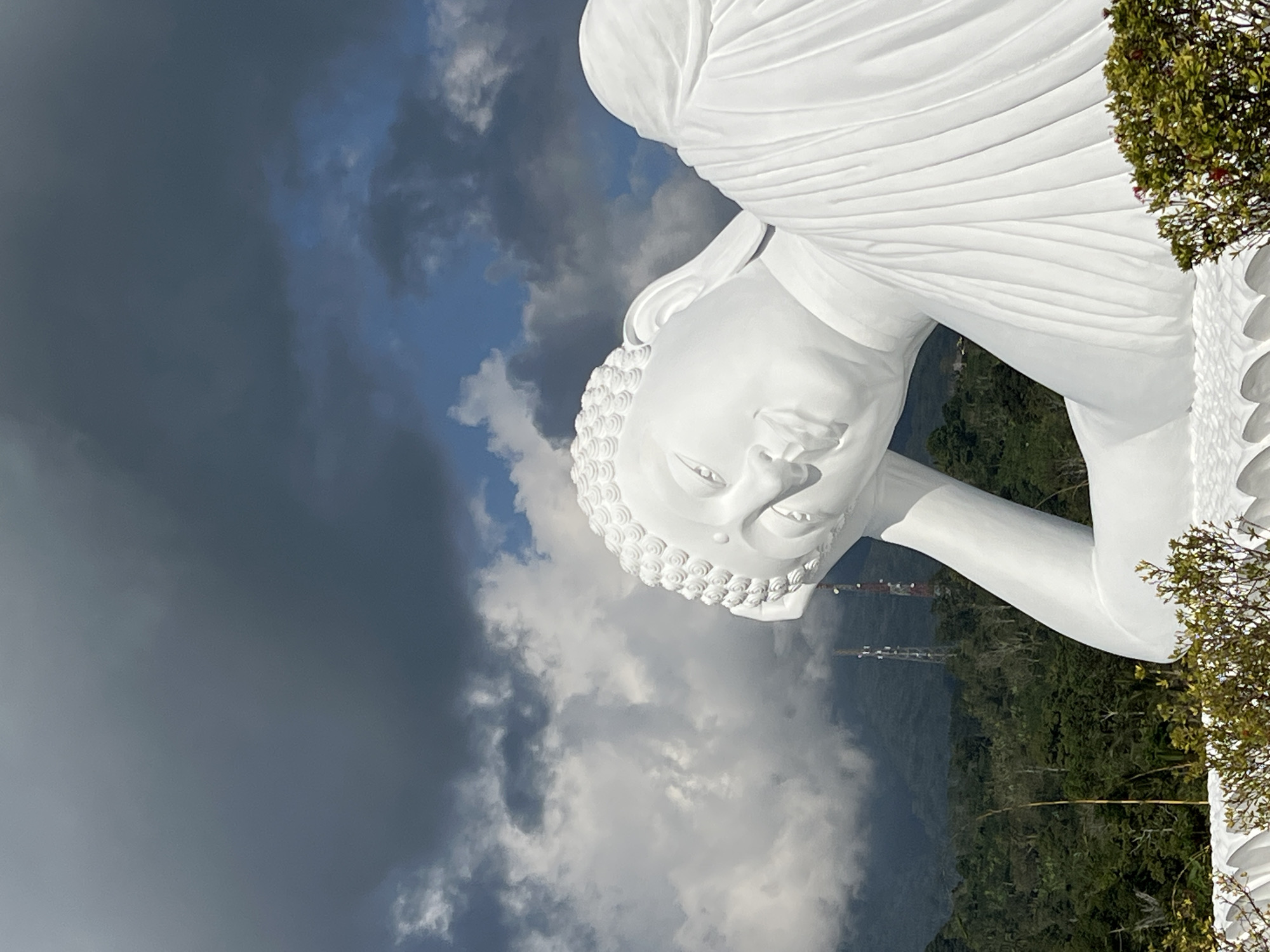 A white, reclining Buddha statue is the primary subject of this outdoor, low-angle photograph. The statue is large and detailed, with its head resting on its arm, and its body draped in flowing robes. The statue is positioned in the foreground and fills a significant portion of the frame.
In the background, a lush green landscape of trees and hills stretches into the distance. Two tall, slender towers with antennas are visible amidst the trees, suggesting a telecommunications or broadcast facility. The sky above is a mix of dark, ominous storm clouds and patches of bright blue sky, with some lighter, wispy clouds interspersed. The overall lighting suggests an overcast day with dramatic cloud formations. The location is identified as Pupuan, Indonesia.