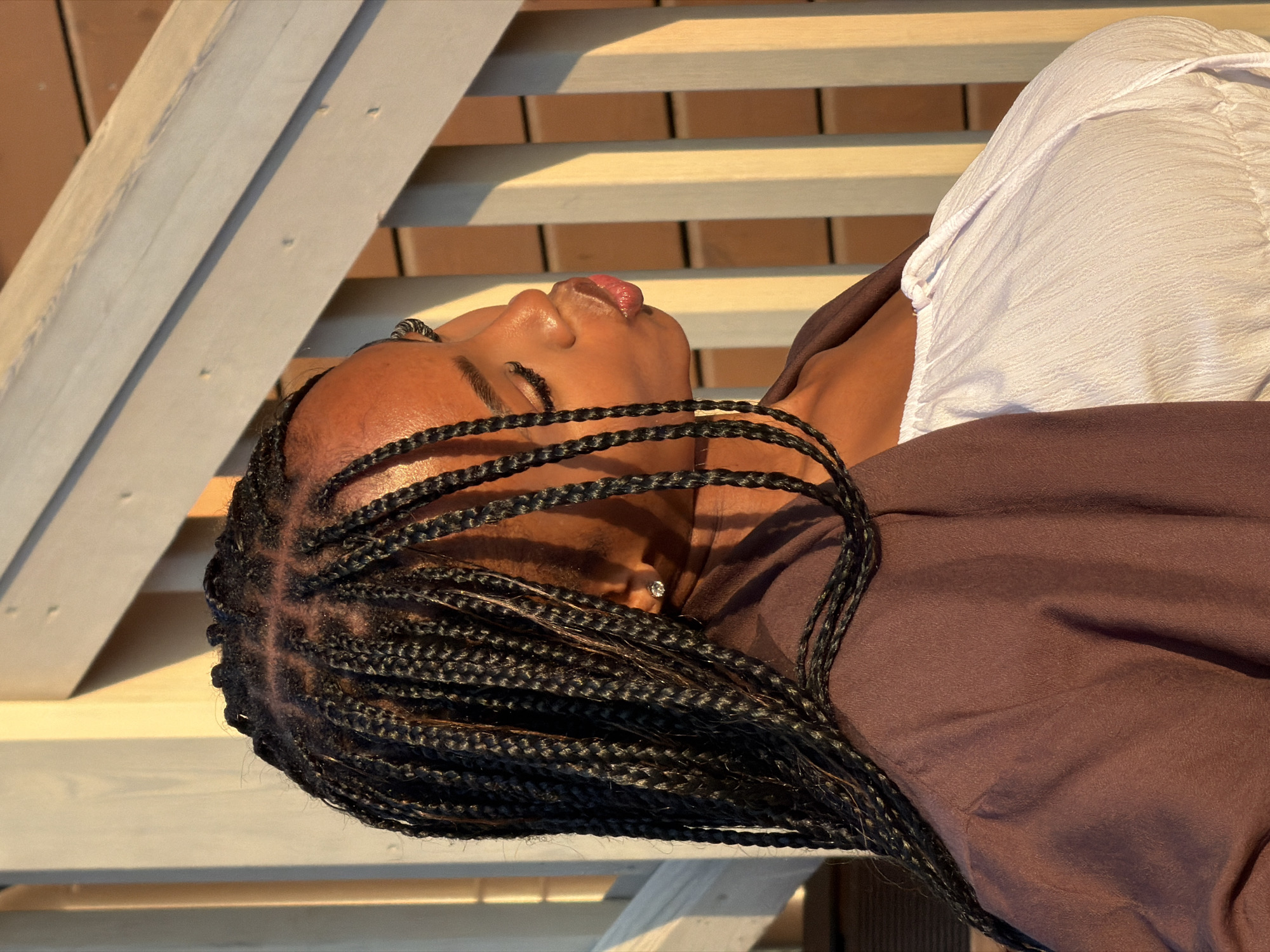 A young Black woman with braided hair is pictured from the chest up, posed in profile outdoors. She is wearing a dark brown jacket over a white textured top with a drawstring. Her eyes are closed, and her lips are puckered as if for a kiss. A diamond stud earring is visible in her left earlobe. The background consists of a wooden deck railing with vertical slats, illuminated by warm sunlight, suggesting a late afternoon or early evening setting. The image does not provide any discernible clues to its specific location within the city of Kazan, Russia, or any other urban environment.