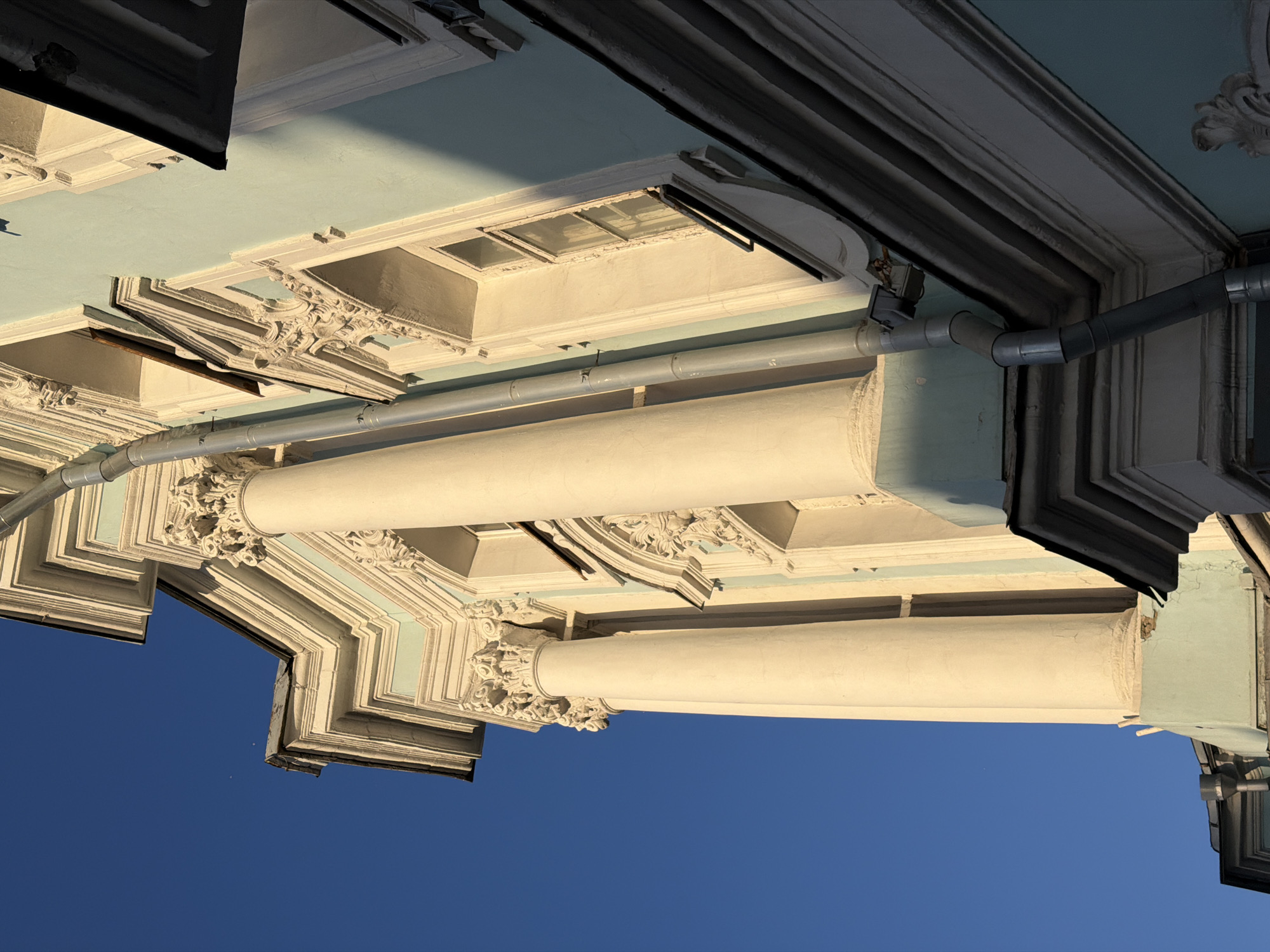 An upward-facing shot shows a building's architectural details against a blue sky, possibly in Moscow, Russia. The building features classical elements, including columns, ornate trim, and possible cornices. A gray pipe runs along the building, adding a modern intrusion into the historical design. The building's underside is painted in light shades, which are contrasted by the dark trim and shadows. The angle suggests a photograph taken while looking up at the building's facade.