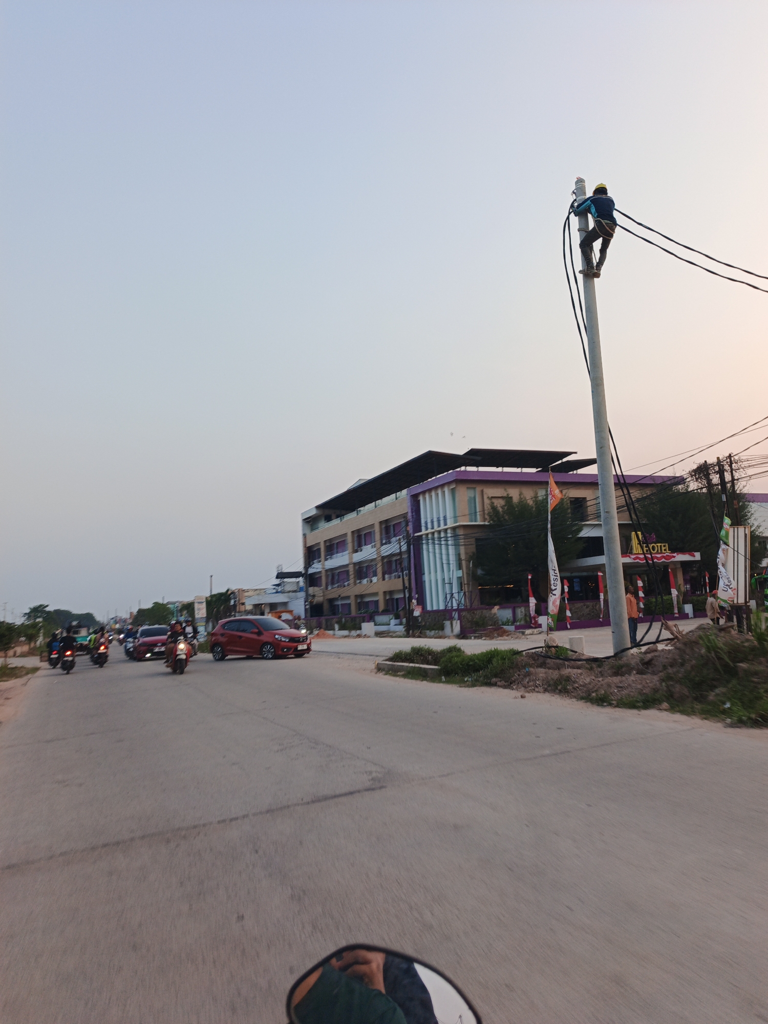 In Batam, Indonesia, a person wearing a yellow hard hat and safety harness works on a utility pole, attending to overhead wires. The scene unfolds on a street with moderate traffic, including motorcycles and cars, under a pale sky. A multi-story building with a distinctive purple trim is visible on the right side of the road, identified by signage as the "ALLE HOTEL". The environment suggests an urban or suburban setting with ongoing development, indicated by dirt mounds and construction debris along the roadside. The perspective is from a moving vehicle, likely a motorcycle, given the reflection in the rearview mirror.