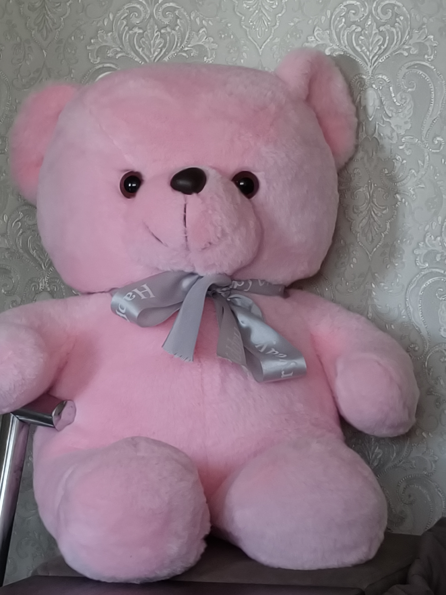 A pink teddy bear with a gray bow sits on a surface. The teddy bear has black eyes and a brown nose. The bow has the word "Happy" printed on it. The background is a light gray wallpaper with a floral pattern. A chrome-colored chair leg is visible in the bottom left corner. The location is Kryvyi Rih, Ukraine.