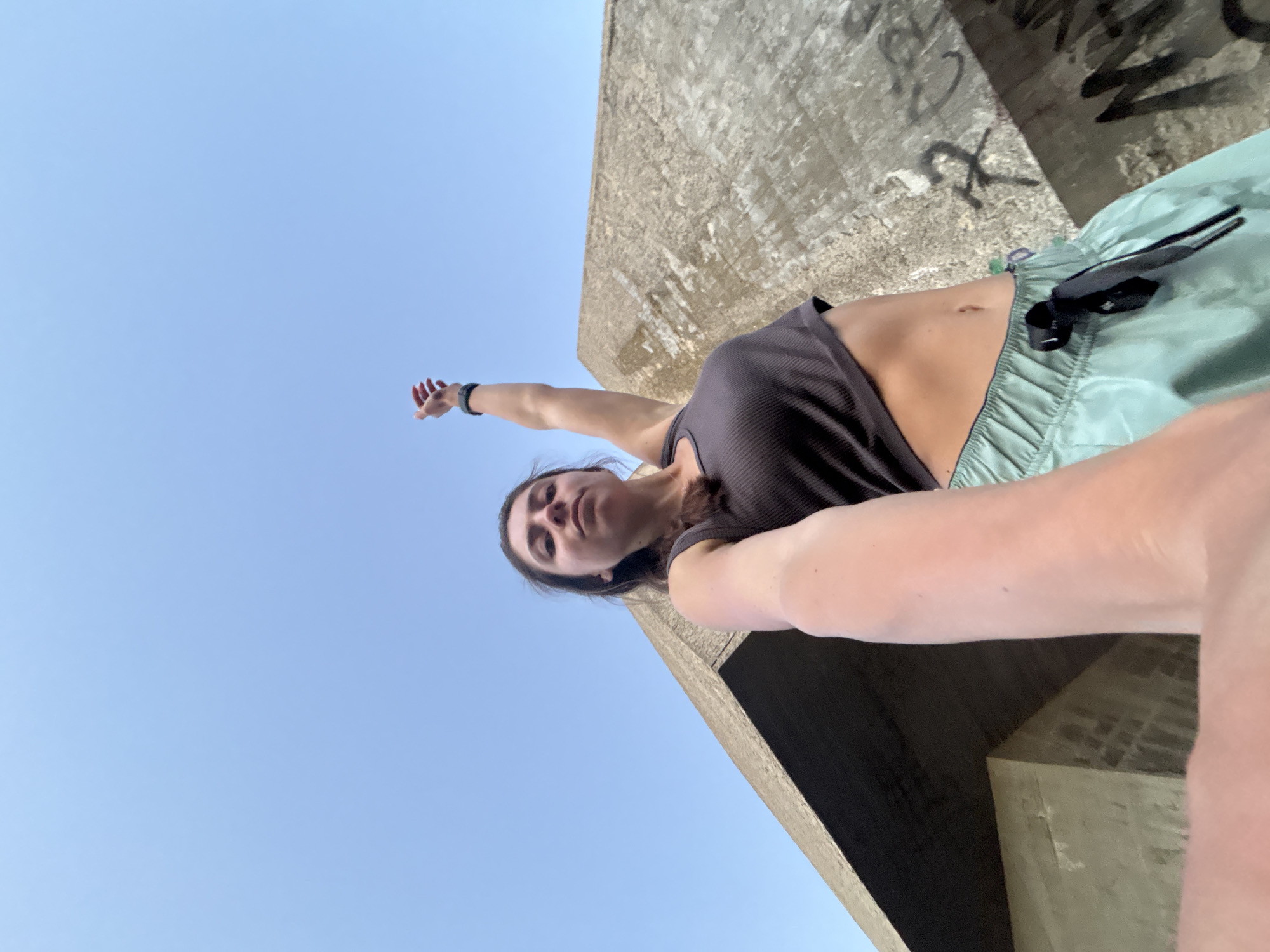 A young woman is taking a selfie in an outdoor urban setting, with a bright blue sky above. She is wearing a dark grey tank top and light blue athletic shorts. Her left arm is raised and extended towards the sky, while her right arm holds the camera. Behind her, a large, angular concrete structure, possibly part of a building or monument, dominates the background. The concrete shows signs of age and weathering, with some graffiti visible on its surface. The overall impression is one of a candid moment captured during an outing, with the concrete architecture suggesting a public space within the city of Varna, Bulgaria.