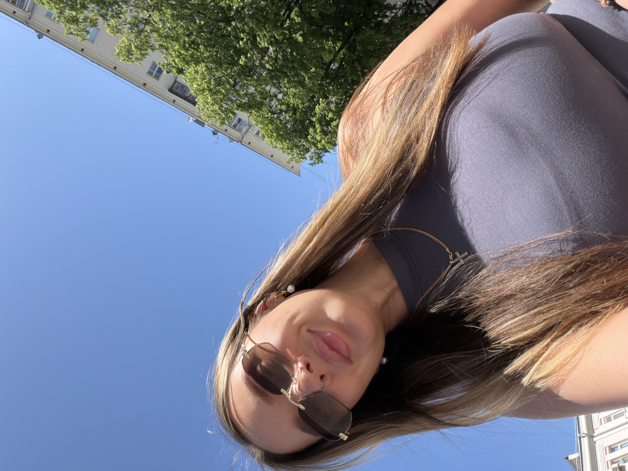 A low-angle shot of a woman outdoors on a sunny day. She is wearing sunglasses and a dark-colored top. A building and a tree are visible in the background. The location is Moscow, Russia, as suggested by the architectural style of the building.