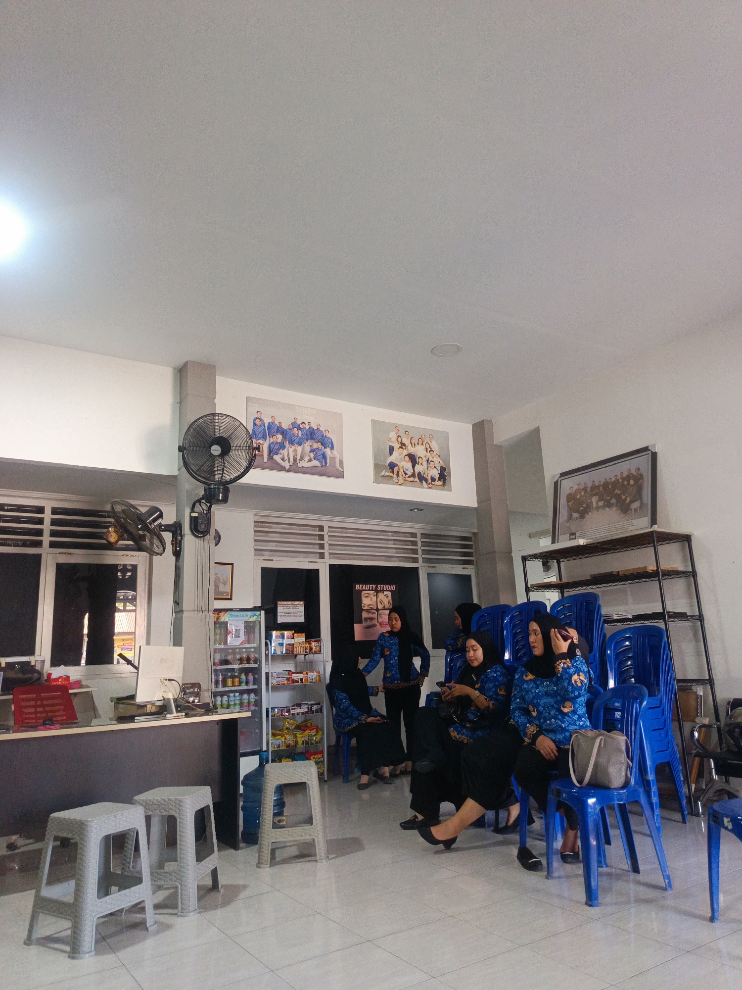 The image depicts the interior of a waiting area, possibly a beauty studio or office, in Gorontalo, Indonesia. Several women, dressed in blue batik patterned shirts and black trousers or skirts, are seated on bright blue plastic chairs. Some are looking at their phones, while others are engaged in conversation. In the background, there are shelves stocked with products, a refrigerator displaying drinks, and framed photographs on the wall. Two large black electric fans are mounted on a pole, and a smaller fan is visible on a desk. The room is well-lit, with white walls and a tiled floor.