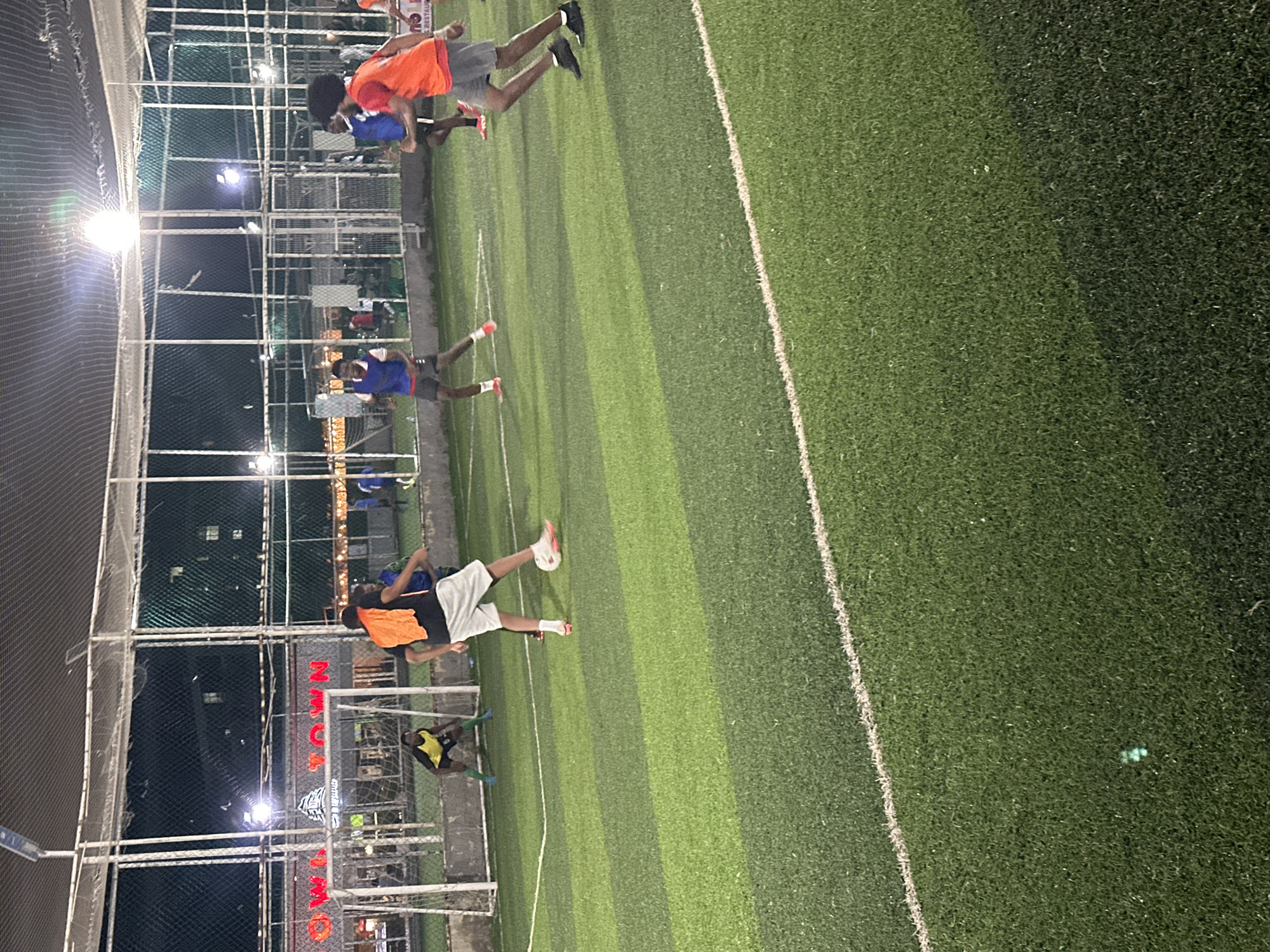 In Abuja, Nigeria, at an outdoor football field with artificial turf, several individuals are engaged in a football match. The field is illuminated by overhead lights, creating a nighttime scene. The players are wearing athletic attire, with some in blue shirts and shorts, and others in orange and yellow jerseys. One player in a yellow jersey is positioned as a goalkeeper in front of a goal. Another player in an orange jersey is kicking a football. The background shows a fence and a building with signage that appears to say "OWN TOWN". The overall environment suggests a recreational or competitive football game taking place within the city.