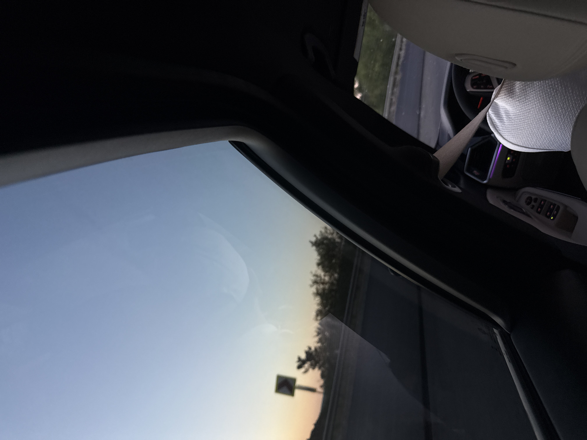 The image shows the interior of a car, likely in motion, with a view through the sunroof and side window. The sky outside is a gradient of light blue to pale orange, suggesting it is either dawn or dusk. A traffic sign, possibly indicating a curve, is visible on the side of the road, along with trees. Inside the car, the steering wheel and dashboard are partially visible. The ambient lighting inside the car appears to be illuminated with purple accents. A person's head and shoulder, wearing a light-colored garment, are visible in the driver's seat, with a seatbelt fastened. The car appears to be on a highway. The location of Fundul Galbenei, Moldova, is mentioned, but there are no specific visual cues in the image to confirm this exact location.