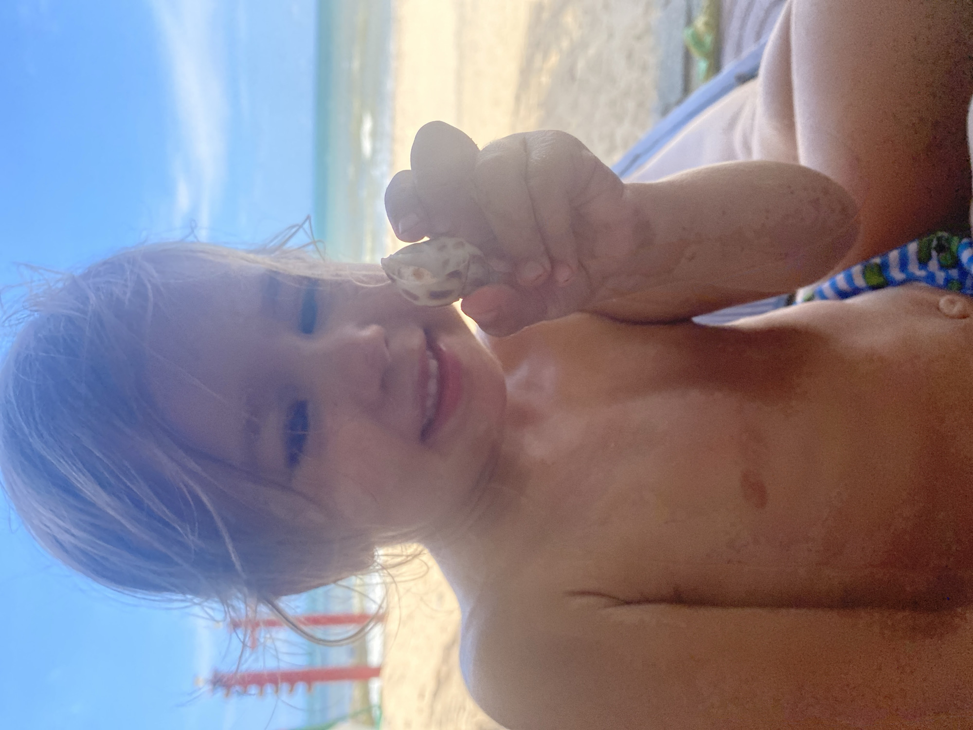 A child, seen from a close-up perspective, is on a beach, holding a small shell up to the camera. The child's face is partially visible, with their mouth slightly open in a smile. Their blonde hair is tousled. The background shows a sandy beach, the ocean, and a clear blue sky with some wispy clouds. The child appears to be wearing a blue and white striped bathing suit. The lighting suggests it is daytime, likely sunny given the beach setting. The overall mood is one of childhood curiosity and enjoyment of a day at the beach.