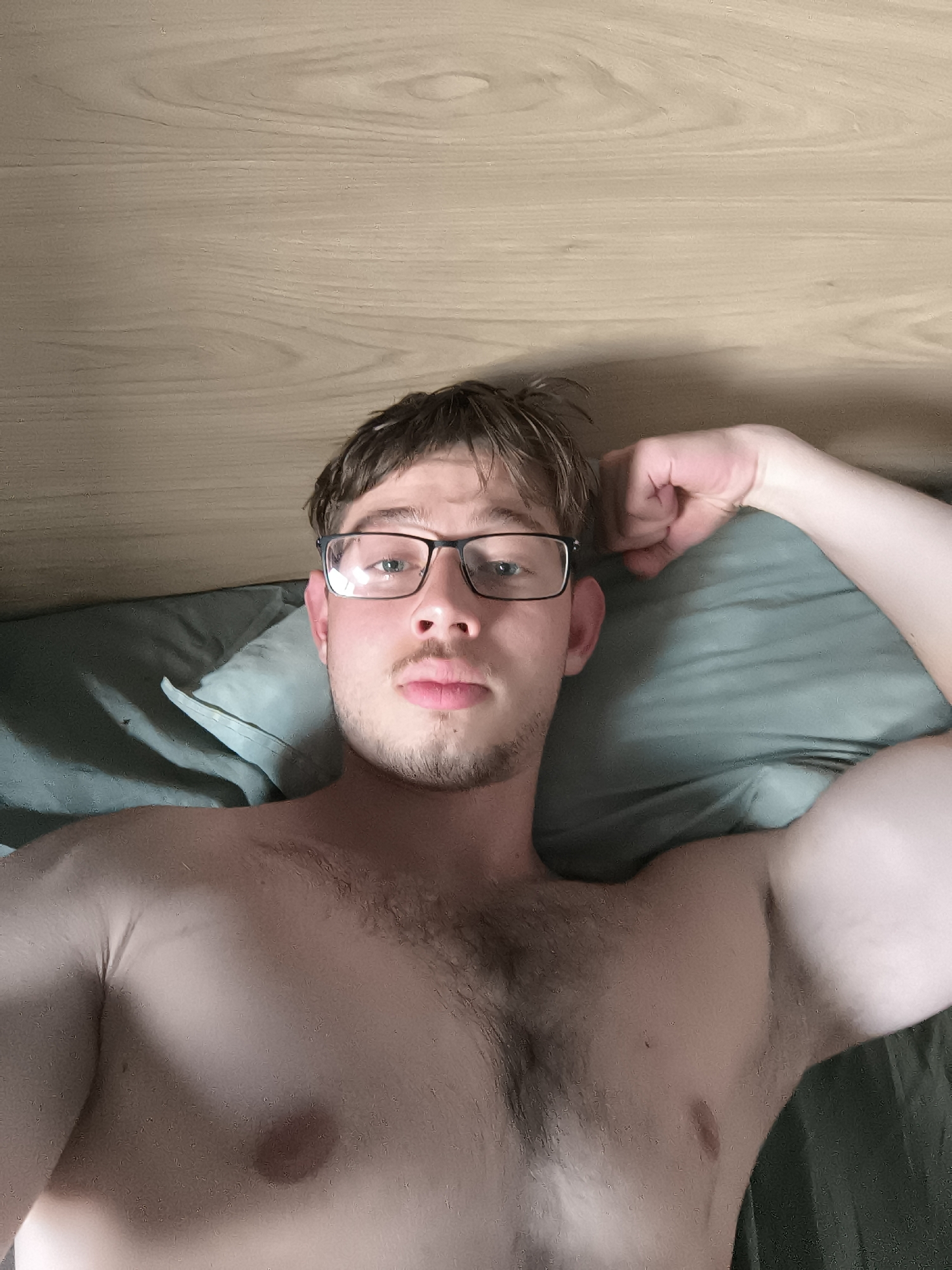 A young man, likely in his late teens or early twenties, is captured in a selfie taken from an overhead perspective, seemingly lying in a bed in an indoor setting in Sofia, Bulgaria. He has short, light brown hair and is wearing black-rimmed glasses. His face shows light stubble or a short beard, and his expression is neutral with slightly pursed lips as he looks directly at the camera. He is shirtless, revealing his upper torso with visible chest hair and a muscular build in his arms. His left arm is bent, with his hand positioned behind his head or flexed. He is resting against a muted teal or green colored pillow and bedding. Behind him, a light wooden panel, possibly a headboard or a wall, is visible. The lighting appears to be ambient indoor light. There is no visible text in the image.