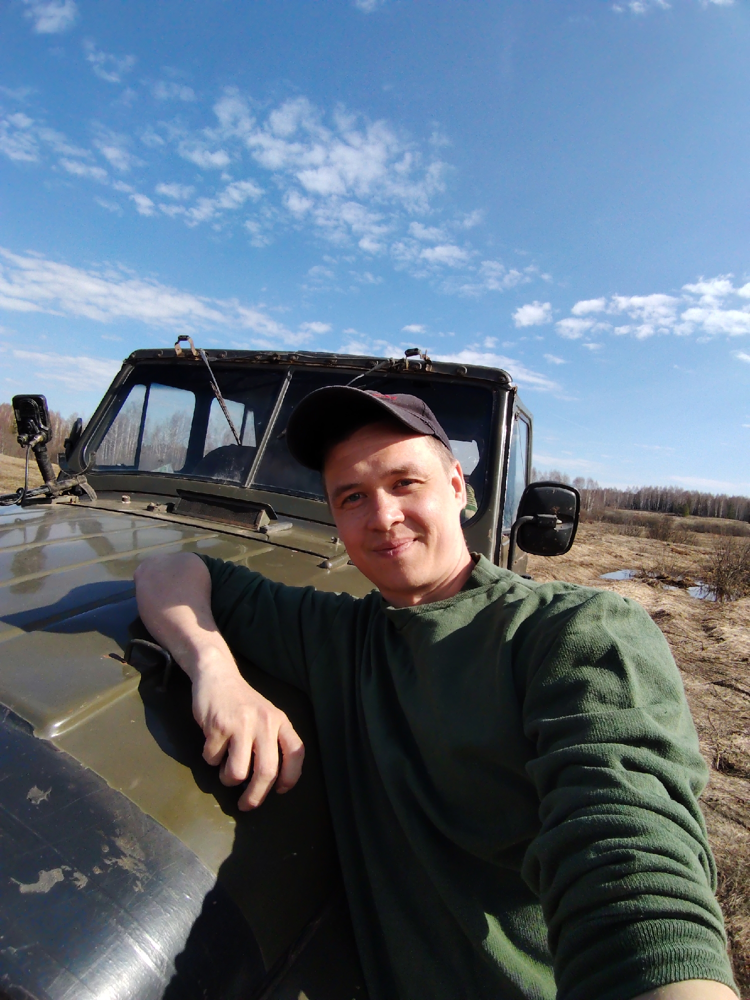 The image is a selfie featuring a man posing outdoors next to a rugged, olive-green utility vehicle, likely in a rural area near Markovskii, Russia. The man is in the foreground, smiling subtly at the camera. He wears a dark baseball cap and a green long-sleeved top, with his left arm resting casually on the vehicle's hood.
The vehicle, which appears to be a military-style off-road truck such as a UAZ, features a prominent upright windshield, side mirrors, and an auxiliary light mounted on the front left. Its sturdy, utilitarian design suggests it is well-suited for challenging terrain.
The background depicts an open, expansive landscape under a bright blue sky with scattered white, puffy clouds. The ground is covered with dry, yellowish-brown grass and sparse vegetation, interspersed with visible puddles of water, suggesting conditions typical of early spring after snowmelt or recent rain. A distant line of bare deciduous trees forms the horizon, reinforcing the early spring or late autumn setting. The sunny weather indicates a clear day. The man's relaxed posture and slight smile convey a sense of contentment or enjoyment of his surroundings. No visible text is present in the image.