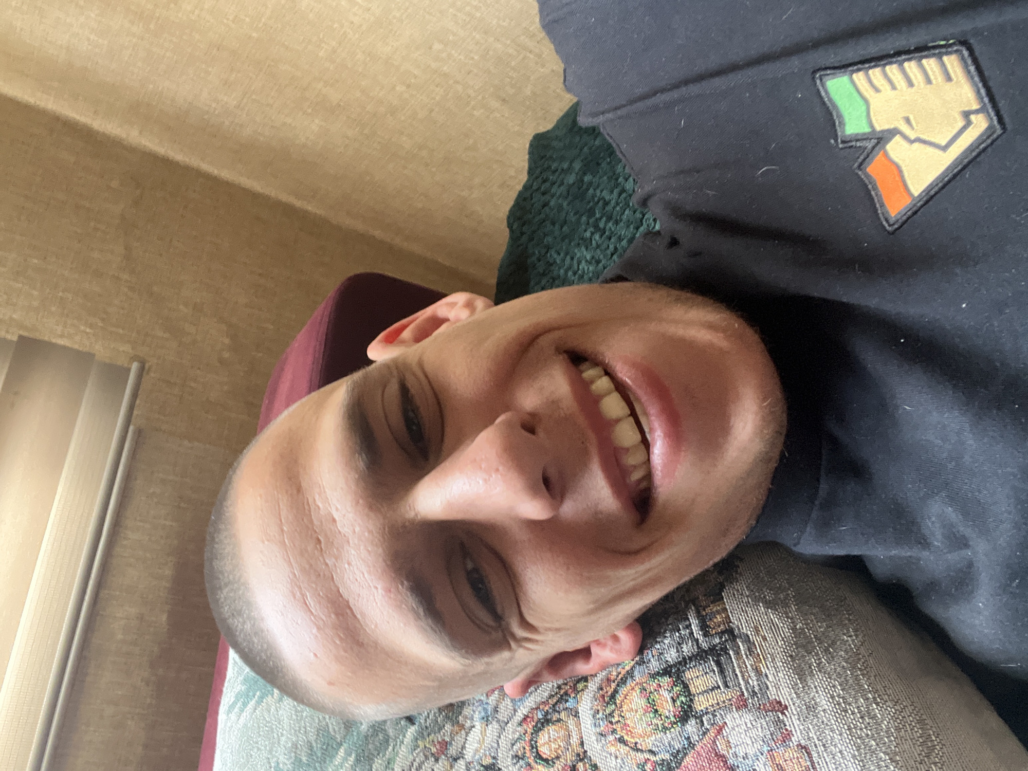 A man with short, light-colored hair is lying down, facing the camera and smiling. He is wearing a black shirt with a patch that appears to be a stylized fist in colors of green, beige and orange. He is lying on a patterned cushion with a floral-type design and maroon-colored fabric. The background includes a textured beige wall and a partially visible window with closed blinds. There is no information to determine if this image was taken in the city of Sofia, Bulgaria.