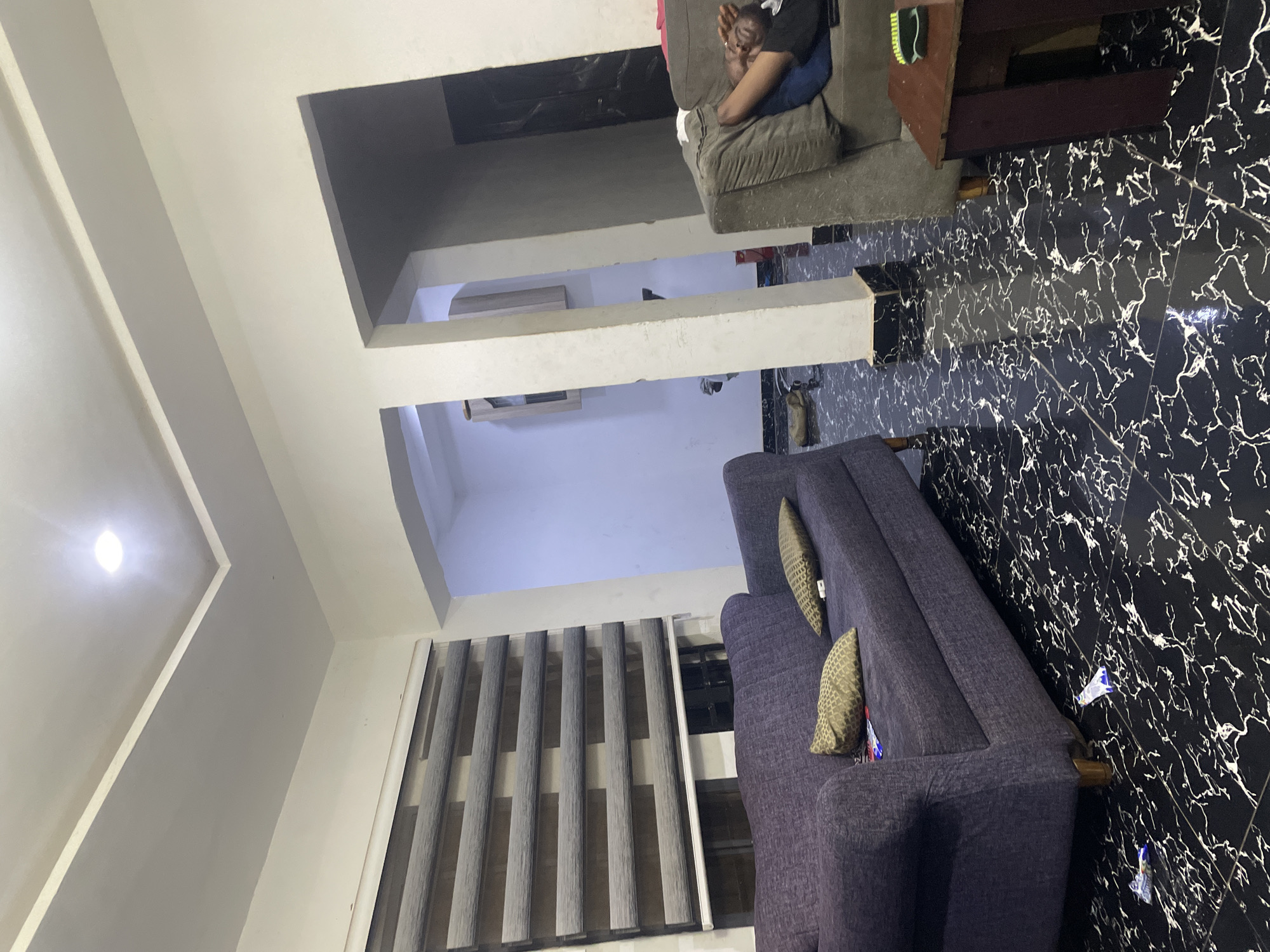 In an indoor living room environment in Abuja, Nigeria, a person is seen sleeping on a grey sofa. The sofa is positioned to the right side of the frame. To the left of the frame, a purple sofa is visible with two decorative pillows on it. The floor is tiled with a black and white marble pattern. In the background, architectural elements suggest an archway leading to another room, which is sparsely furnished and dimly lit. The lighting in the room is primarily from an overhead fixture.