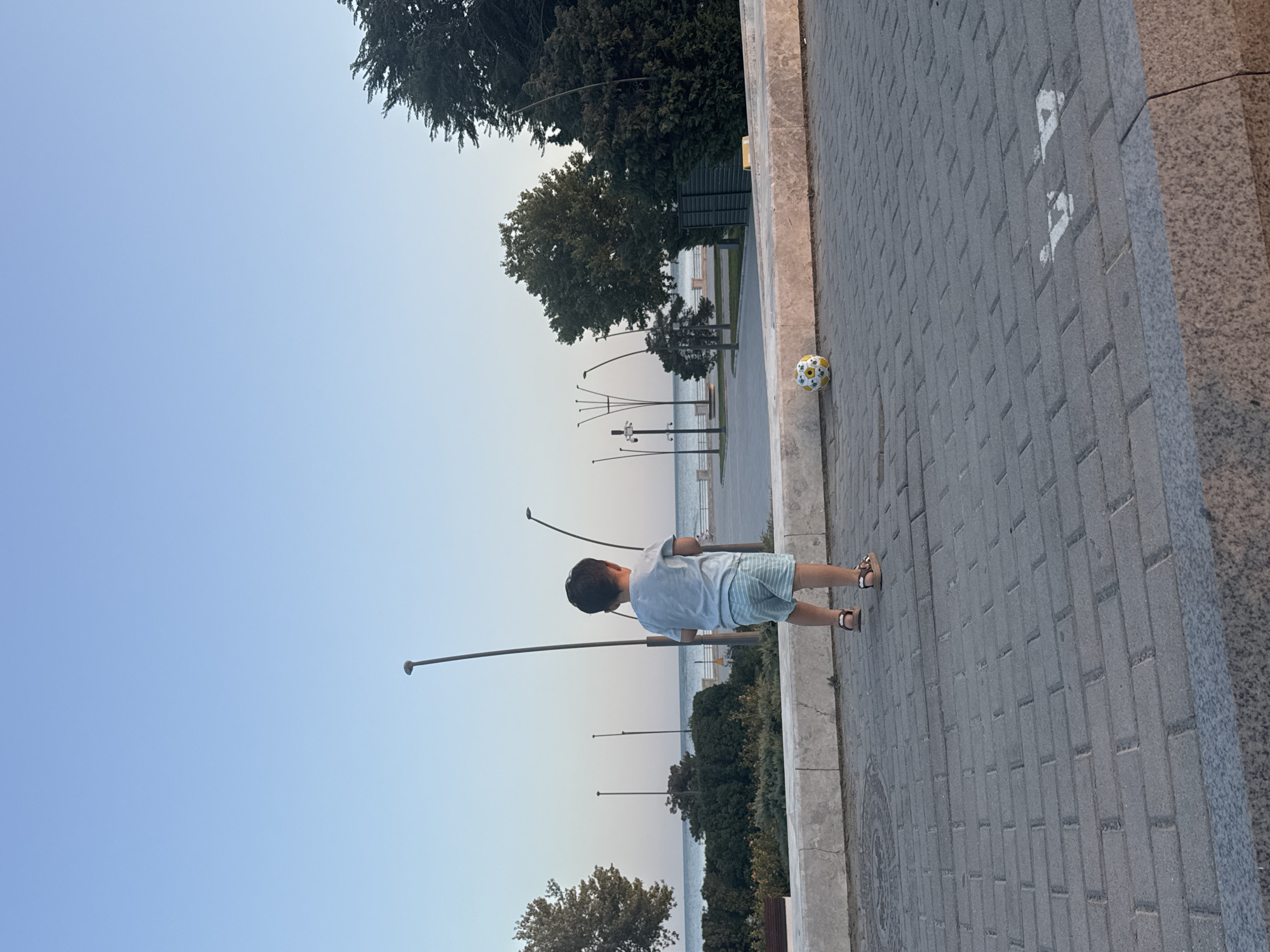 A young child, seen from behind, stands on a paved promenade overlooking the Caspian Sea. The child is wearing a light blue t-shirt and striped shorts, and brown sandals. A soccer ball with yellow and white markings rests on the ground to the child's right. In the background, the calm sea stretches to the horizon under a clear blue sky. Several lampposts line the promenade, and lush green trees and bushes are visible along the coastline. The setting appears to be a public park or a beachside walkway in Khatay Raion, Azerbaijan, given the context. The overall atmosphere is peaceful and idyllic.