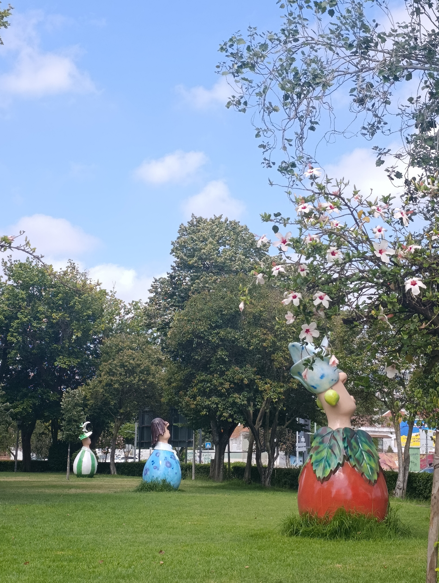 The image captures a sunny day in a park, likely in Setúbal, Portugal, with a bright blue sky dotted with fluffy white clouds. In the foreground, a lush green lawn is adorned with three whimsical, larger-than-life sculptures. To the right, a prominent sculpture resembles a red apple with green leaves, topped by a light blue head with a curious expression, holding what appears to be a bird-shaped pitcher and two lime-green spheres. To the left, two other sculptures stand out: one is a rounded figure wearing a blue dress with darker blue polka dots and a dark brown wig, and the other is a vertically striped green and white figure wearing a green hat with a crescent moon adornment. Behind the sculptures, a line of mature trees with dense green foliage creates a natural backdrop. In the distance, beyond the trees, buildings and streetlights are partially visible, suggesting an urban setting. A flowering tree with delicate white and pink blossoms hangs over the scene on the right, adding a touch of natural beauty. The overall atmosphere is one of playful artistry and serene parkland.