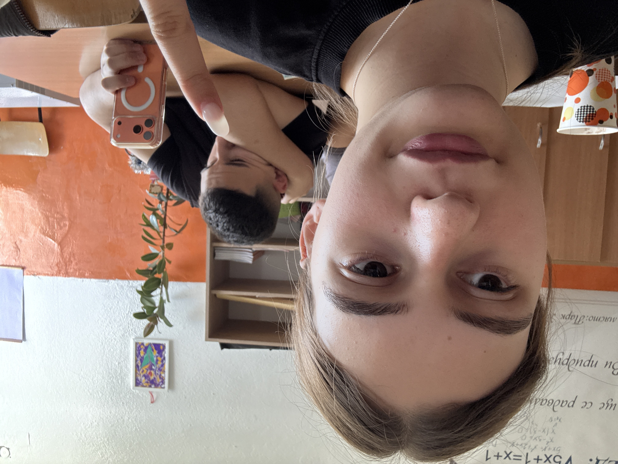 The image is an upside-down selfie taken indoors, possibly in a classroom. The camera is close to the face of a young woman with light skin, light brown hair, and a silver necklace. Her face fills the lower half of the frame. Behind her and to the left, a young man with short, dark hair is resting his head on his arms at a desk. His face is partially obscured. A hand holding a phone with a unique circular design on the back is visible to the left of his head.
The background includes an orange and white painted wall, a small framed artwork hanging on the wall, a green plant, and a wooden shelving unit. To the right, there's a whiteboard or paper with handwritten text in a foreign language. The text contains the words "място: Парк" which translates to "place: Park", but there is no direct indication of the city or country within the scene. However, since the OCR includes Bulgarian characters, it strongly suggests the scene is in Bulgaria. The handwriting includes the word "Парк" which would likely refer to a park nearby.