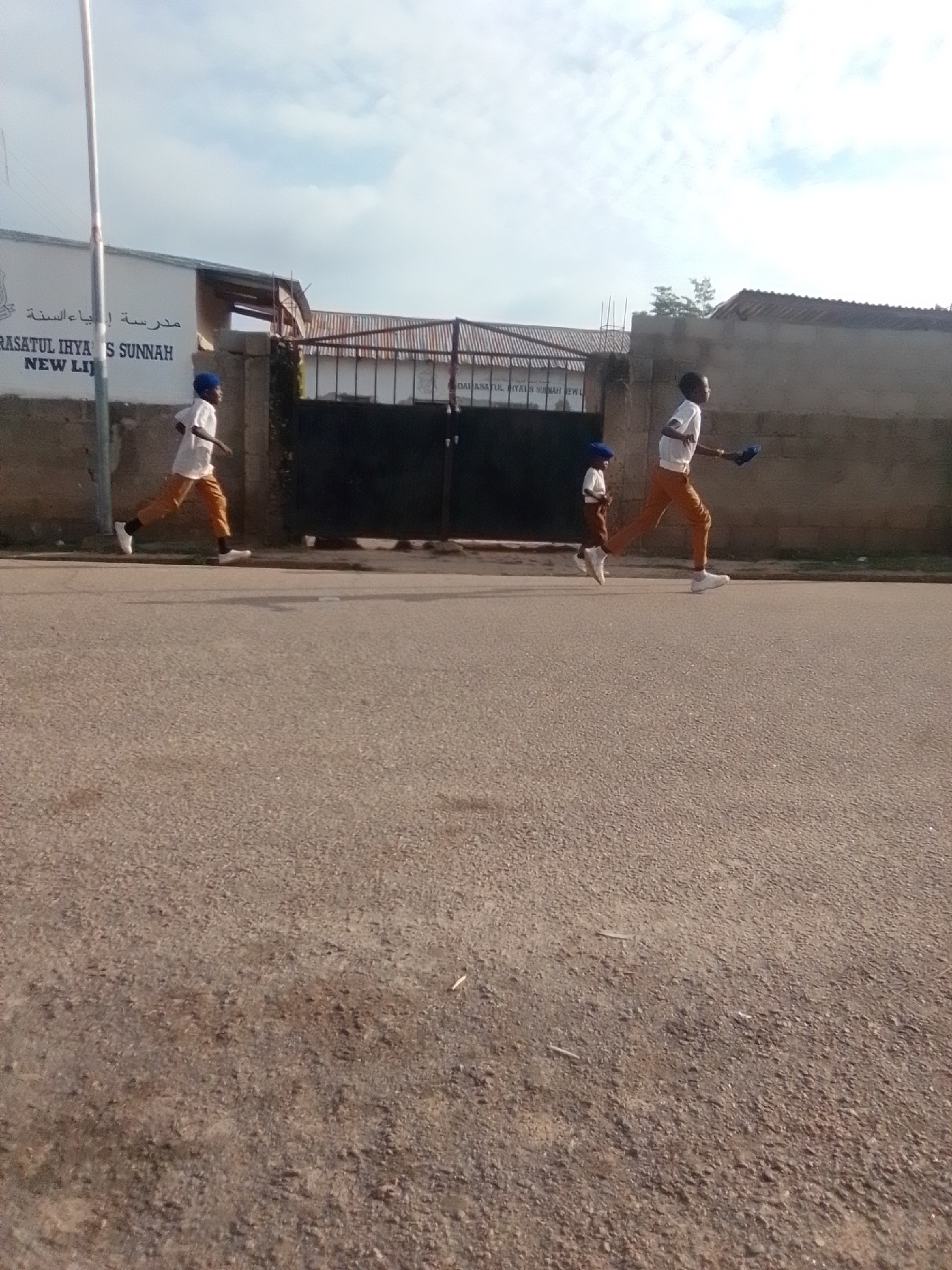 The image depicts a street scene in Liji Hill, Nigeria, with three individuals in the foreground. Two children are running, one slightly ahead of the other. The child in the lead, wearing a white short-sleeved shirt and light brown pants, is captured mid-stride, with their right leg extended forward and left leg trailing. They have a dark blue object in their right hand. The second child, wearing a similar white shirt and brown pants, with a blue cap, is positioned behind and to the left of the first. They are also running, with their body turned slightly towards the viewer. Further behind and to the left, a third child, dressed in a white long-sleeved shirt and light brown pants, is also running. They appear to be wearing a blue cap. The background features a building with Arabic script on its facade, possibly a school, and a dark metal gate. The ground is paved with asphalt. The sky is cloudy, with a bright sun illuminating the scene.