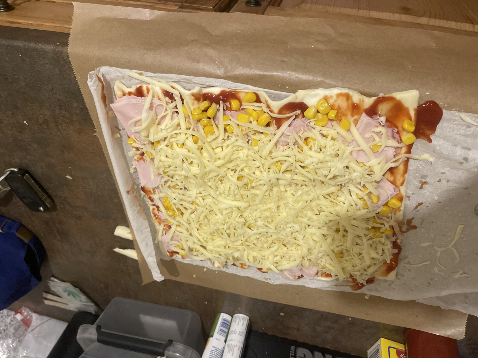 A high-angle, full shot reveals a partially assembled pizza on baking paper, presumably on a countertop. The pizza is rectangular and features a layer of shredded cheese covering most of the surface. Beneath the cheese, slices of ham, corn kernels, and red sauce are visible. The baking paper extends beyond the pizza and is crumpled in places. In the lower left corner, there's a blue bag and a black container with a translucent grey lid. To the right of these items, there are two cylindrical containers with printed labels and a black object. Part of a yellow carton with the letter "K" on it is visible in the lower right corner. The environment suggests an indoor setting, possibly a kitchen.