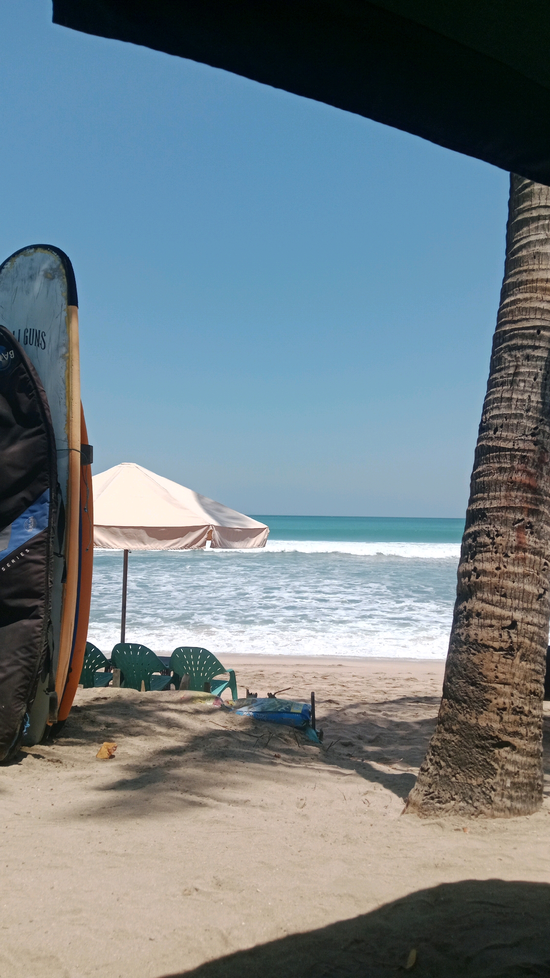 This is an eye-level, medium shot of a beach in Kuta, Indonesia, on a clear, sunny day. The foreground is dominated by a sand-covered ground, with the textured trunk of a palm tree on the right side of the frame. In the mid-ground, there are several green plastic beach chairs, a beige beach umbrella providing shade, and two surfboards leaning against each other. Beyond that, the ocean stretches out with white waves breaking on the shore, and the horizon is marked by a clear blue sky. The image appears to be taken from under a dark structure, possibly a roof or awning, which casts a shadow across the sand in the foreground. There are no people visible in the frame.