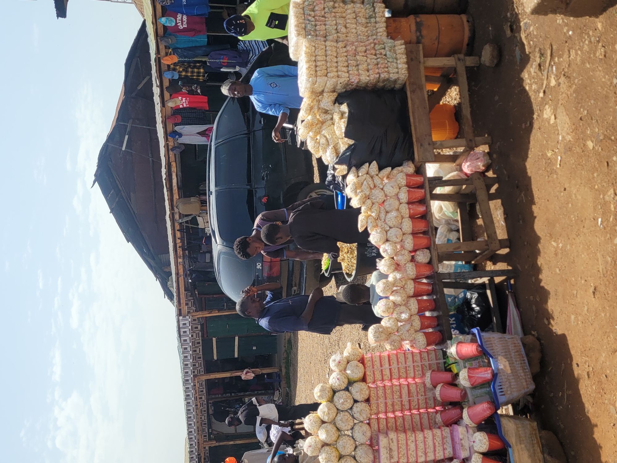 The image depicts an outdoor market scene in Jos, Nigeria, likely a roadside stall. In the foreground, several stalls are laden with bags and cups of popcorn. The popcorn is packaged in clear plastic bags, with some stacked in large quantities and others in smaller red cups. To the left, there are pink rectangular containers filled with popcorn.
A group of men are gathered around one of the stalls. One man in a blue t-shirt is interacting with a man in a light blue traditional outfit. Another man in a yellow long-sleeved shirt with a black patch on the chest stands to the right, also near the popcorn. Behind them, a dark-colored SUV is parked.
In the background, a row of open-fronted stalls selling clothing is visible. T-shirts and jackets are hung on display. The structures appear to be made of wood and corrugated metal, with a somewhat weathered appearance. The ground is dusty and appears to be unpaved, typical of a market environment. The sky is visible, with some clouds. The overall scene suggests a busy commercial activity in an open-air market.