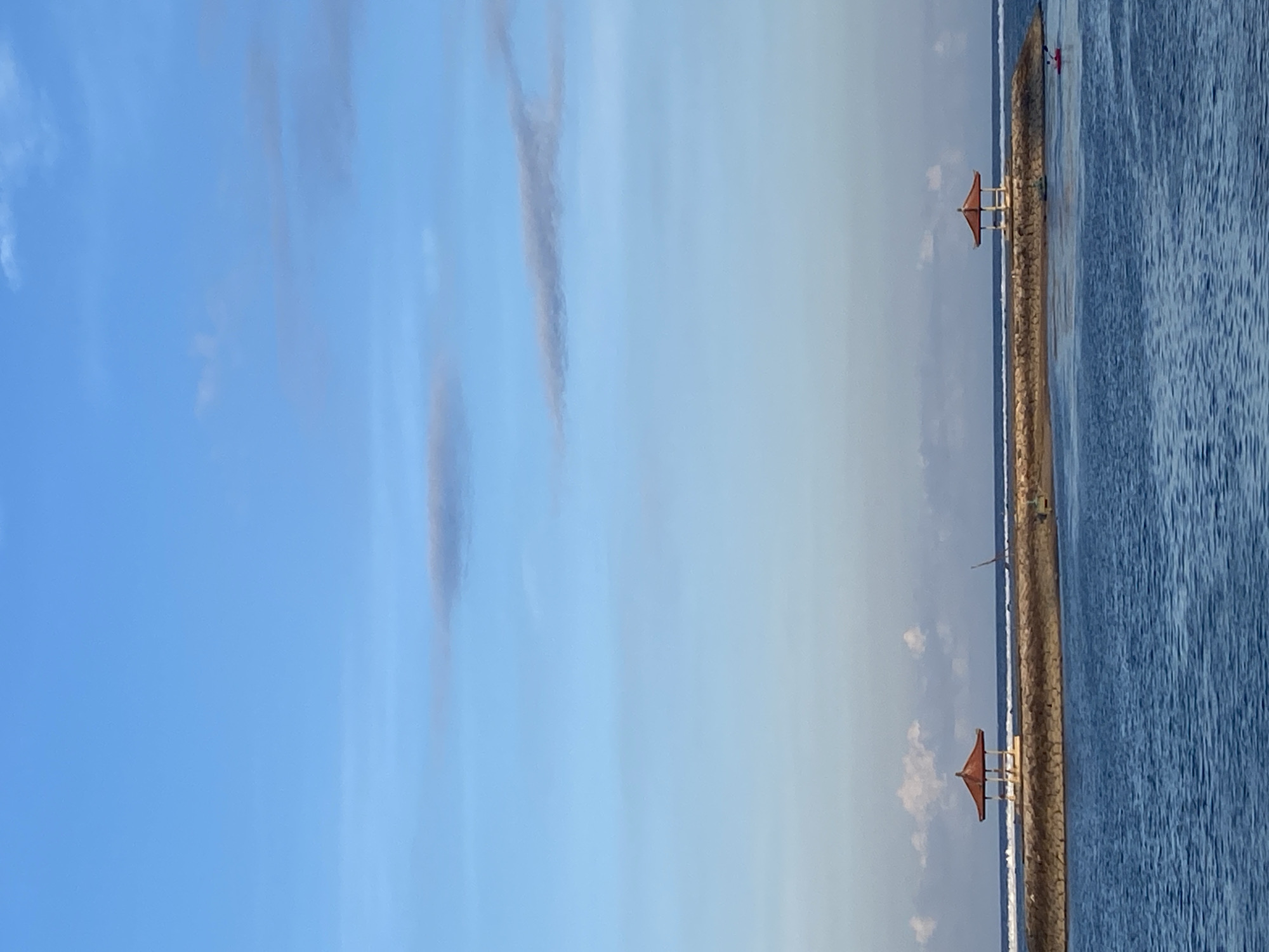 The image depicts a serene seascape at Pantai Sanur, a beach located in Denpasar, Indonesia. In the foreground, deep blue ocean water stretches towards a stone jetty. The jetty is adorned with two traditional Balinese gazebos, each with a red, tiered roof and open sides, providing a picturesque view of the sea. Waves gently break along the shore behind the jetty. In the distance, a single individual can be seen paddling a red kayak on the water, adding a subtle element of human activity to the tranquil scene. The sky above is a gradient of light blue, with scattered, wispy clouds, suggesting a clear and calm day.