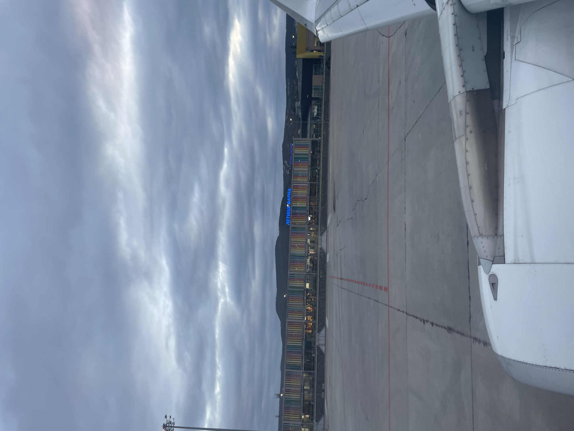 The image shows the Varna Airport (VAR) in Aksakovo, Bulgaria, as seen from the window of an airplane. The airport terminal building is visible, with its colorful facade and the "ЛЕТИЩЕ ВАРНА" sign illuminated in blue. The airplane wing and engine are in the foreground. The airport tarmac and distant hills are also visible under a cloudy sky.