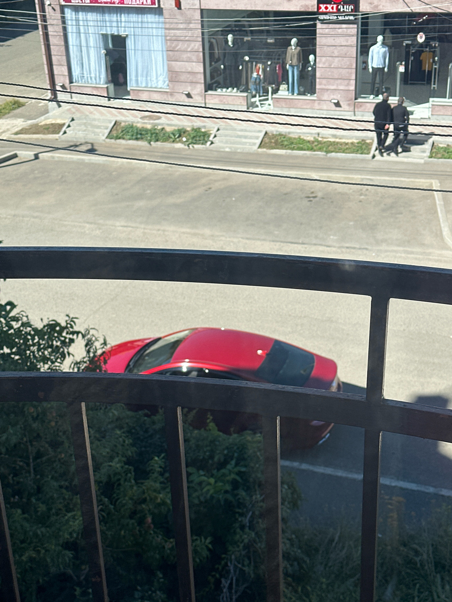 A view from a balcony in Gyumri, Armenia, looks down upon a street and a row of shops. A bright red car is parked on the street, its rear end facing the camera. In front of the shops, two men in dark clothing walk away from the camera, descending a set of stairs. The shops have large windows displaying mannequins dressed in clothing. One shop appears to be a flower shop with light blue curtains visible through its doorway. The overall scene is set on a sunny day, with shadows cast by the buildings and a metal railing in the foreground.